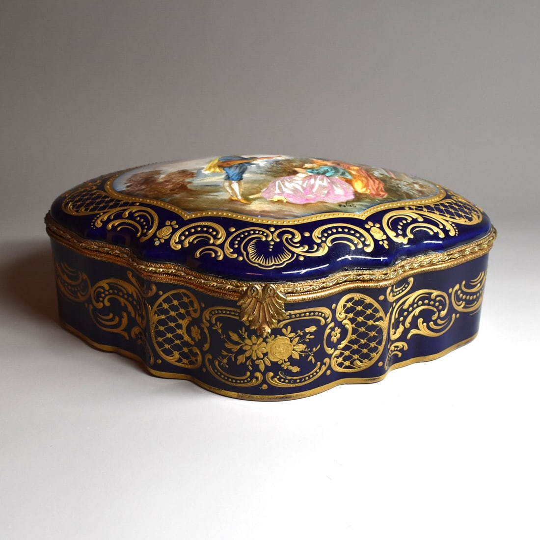 A large SEVRES-style porcelain dresser box: A large antique Sevres style porcelain brass mounted hinged jewelry casket / jewel box of cartouche shape with cobalt-blue ground, the cover painted with a catrtouche featuring an 18th century style b