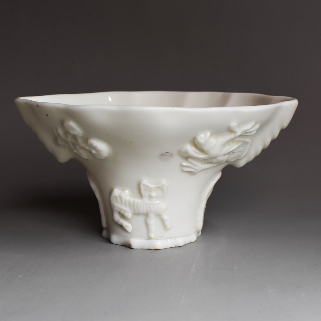 An antique Chinese Dehua porcelain libation cup, Qing Dynasty, 17th century: An antique Chinese Dehua / blanc-de-Chine porcelain 'Rhinoceros-horn-form' libation cup. The vessel is molded and applied with figures of deer, beasts or crane beneath flowering branches and a dragon