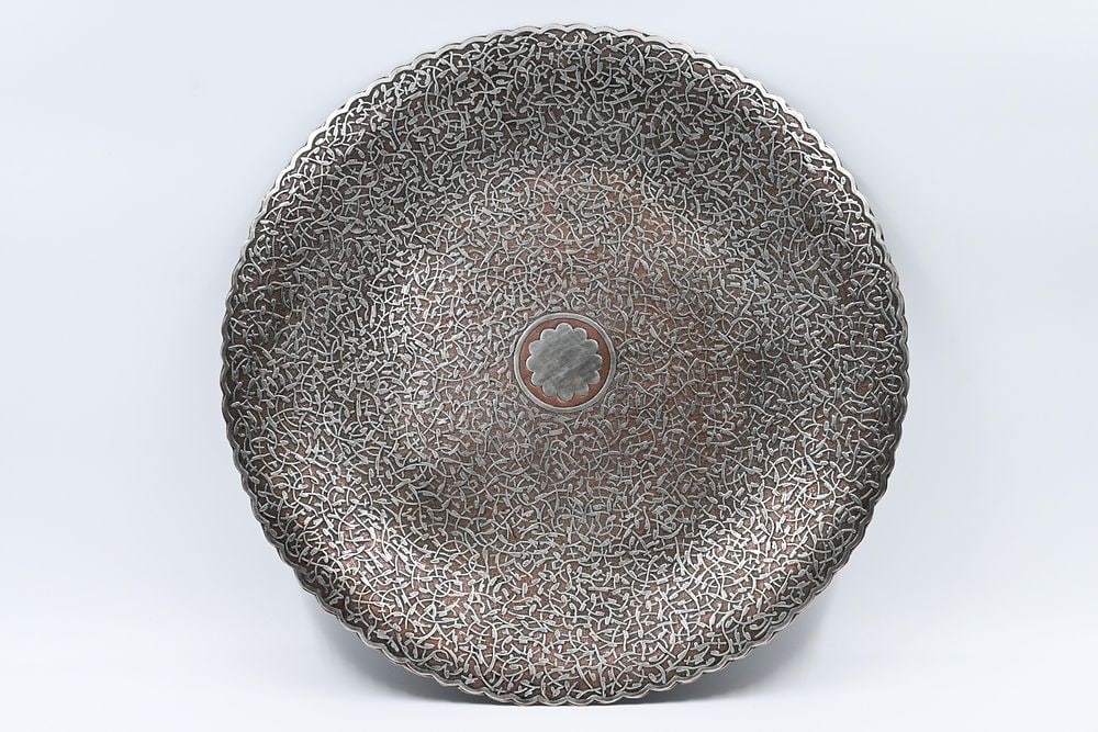A large plate made of brass with damask work decorated with damask work with silver, brass, copper: A large plate made of brass with damask work decorated with damask work with silver, brass, copper inlays and inlays combined with arabesques and calligraphic motifs. Dimensions: diameter 34.7 cm. Sou