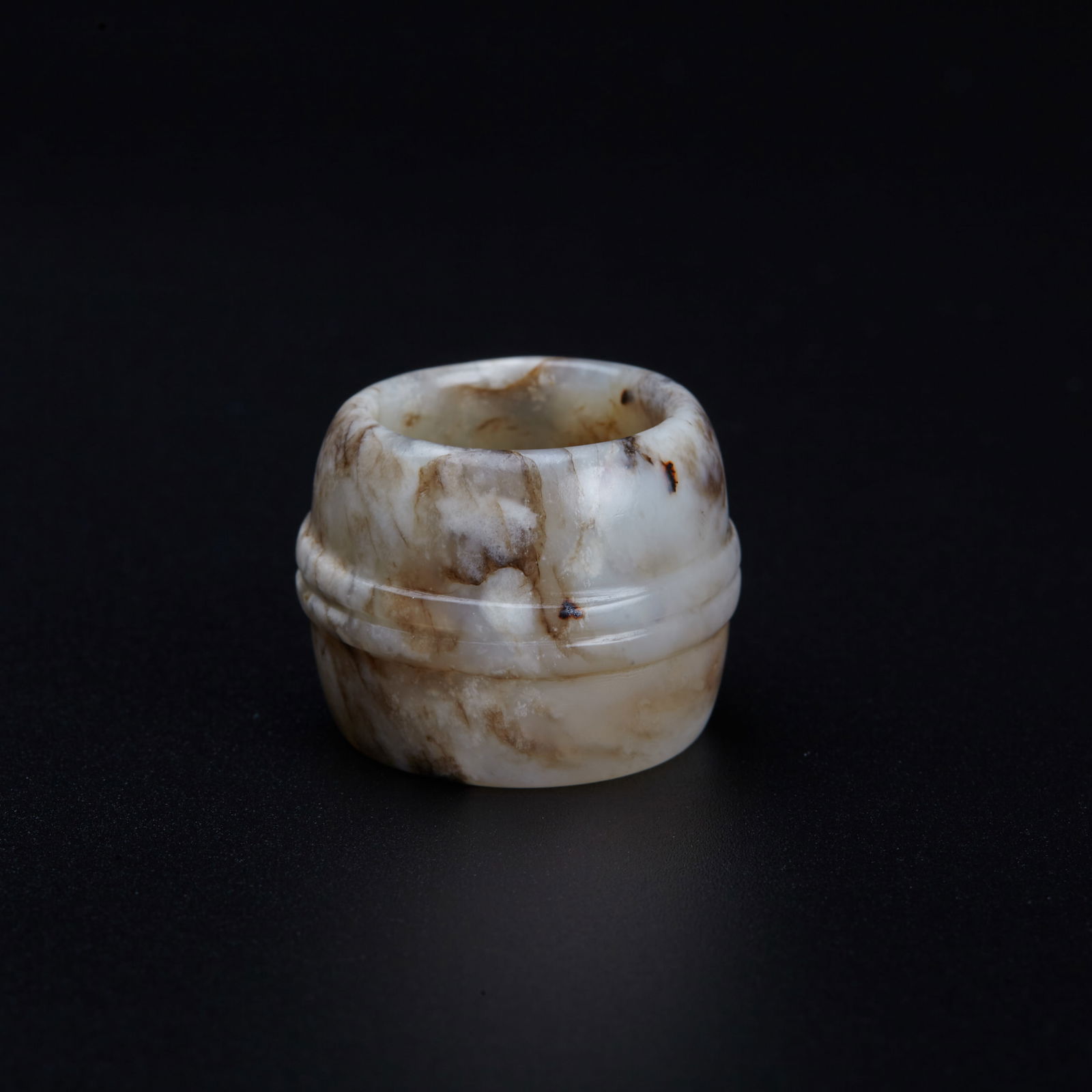 A white jade thumb ring, Qing Dynasty (Bonhams London): A white jade thumb ring, Qing Dynasty (Bonhams London) a mutton fat and frey barrel-shaped ring, the stone of light cream colour with variegated inclusions Provenance: Bonhams London Grosvenor House
