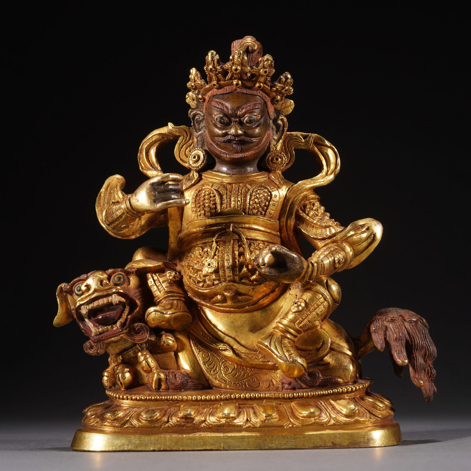 A SINO-TIBETAN BRONZE-GILT GOD OF WEALTH: A SINO-TIBETAN BRONZE-GILT GOD OF WEALTH Casting seated on a recumbent lion above a lotus base, the left hand raised holding a mongoose, the figure depicted clad in an armour, the stern face flanked b