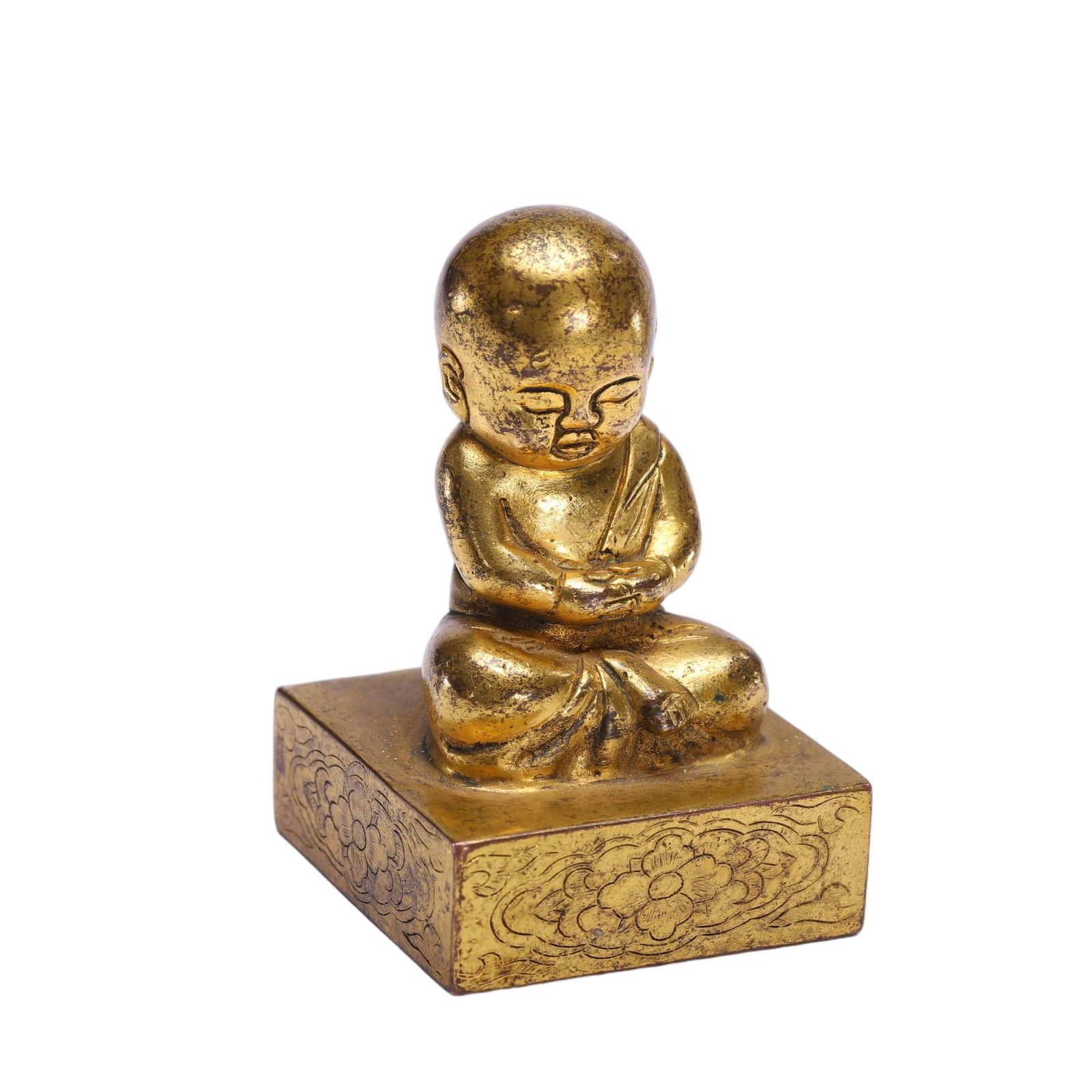A BRONZE-GILT MONK SEAL: A BRONZE-GILT MONK SEAL This refined Chinese seal is cast from gilt-bronze, featuring a square base intricately incised with scrolling floral patterns. Atop the base, a plump boy is depicted seated