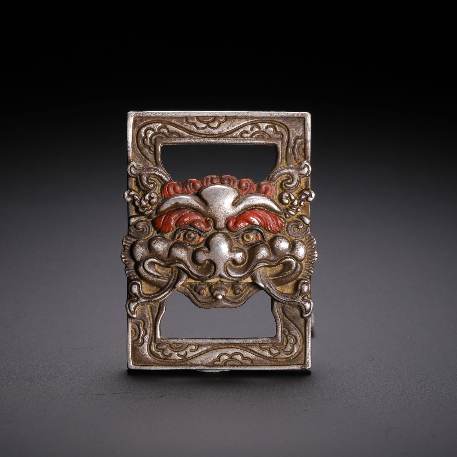 A COPPER ALLOY SUTRA BELTHOOK: A COPPER ALLOY SUTRA BELTHOOK This striking Chinese copper ritual object is cast in the form of a square frame, centered by a fierce, stylized beast mask. The mask features vividly painted red eyebrow