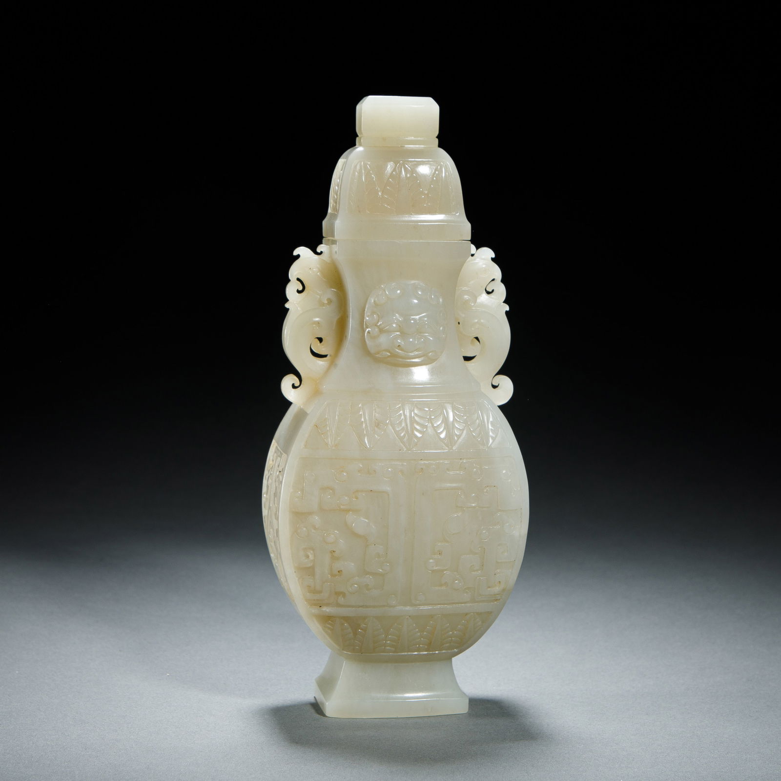 A CHINESE ARCHAISTIC CARVED WHITE JADE VASE WITH COVER: A CHINESE ARCHAISTIC CARVED WHITE JADE VASE WITH COVER This impressive covered vase is carved from a single piece of luminous, pale celadon Hetian jade. It features a flattened, pear-shaped body decor