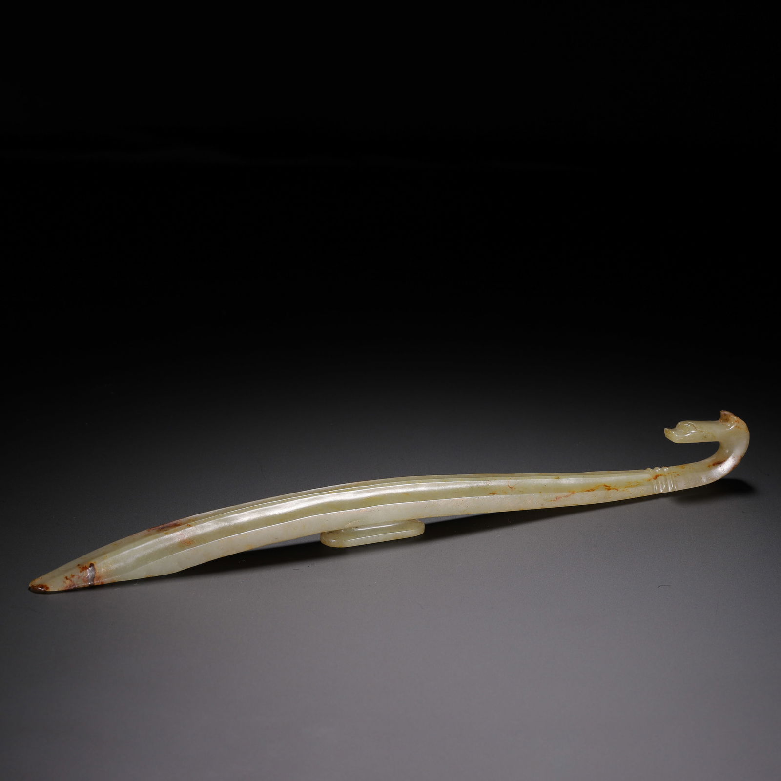 A CARVED WHITE JADE BELTHOOK: A CARVED WHITE JADE BELTHOOK This elegant belt hook is carved from pale, translucent Hetian jade, with natural russet inclusions concentrated at the tip and along the curved terminal. It has a slender