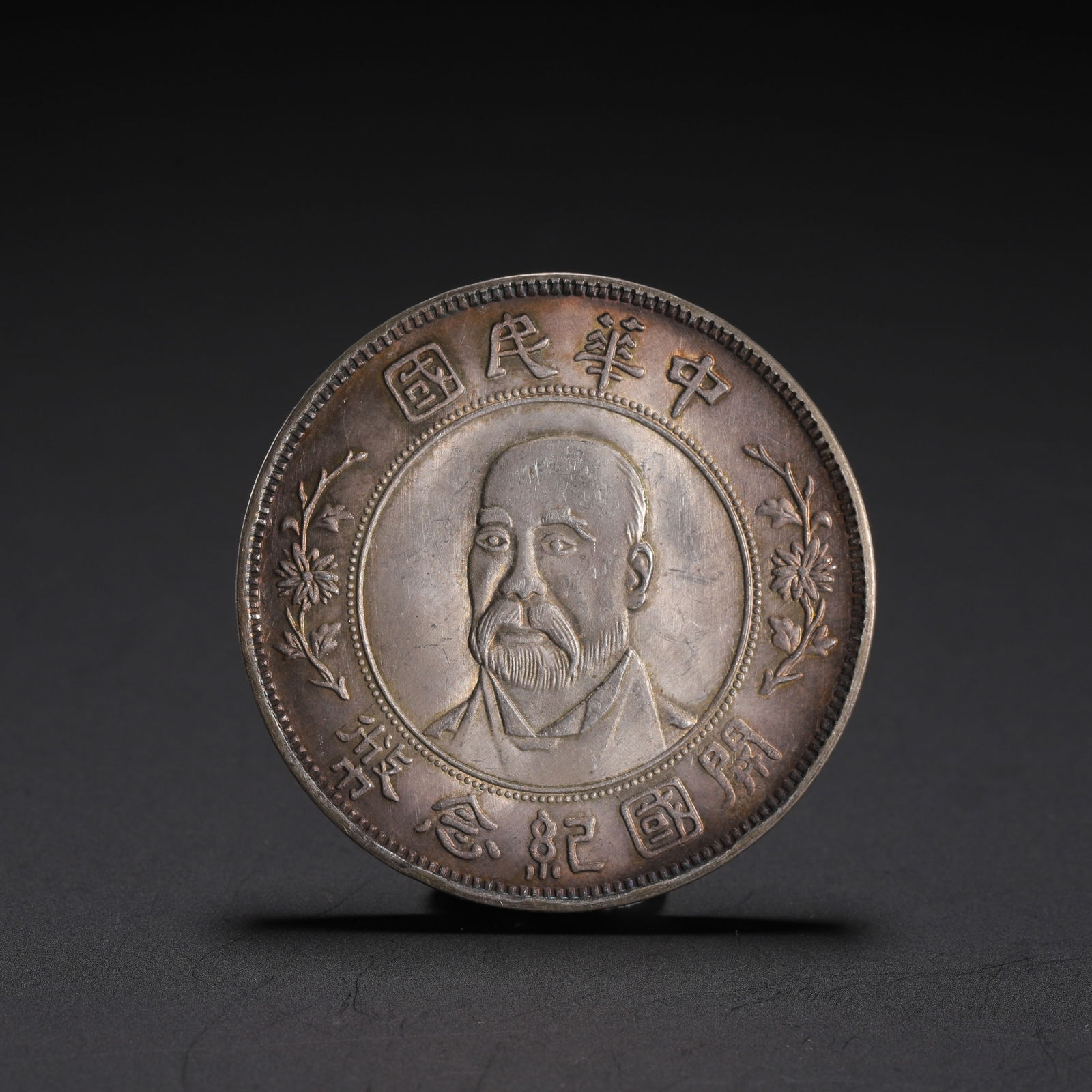 A CHINESE SILVER COIN: A CHINESE SILVER COIN This silver coin is struck with a central portrait of a bearded figure, encircled by the characters “??????????” (Republic of China Founding Commemorative Coin). The