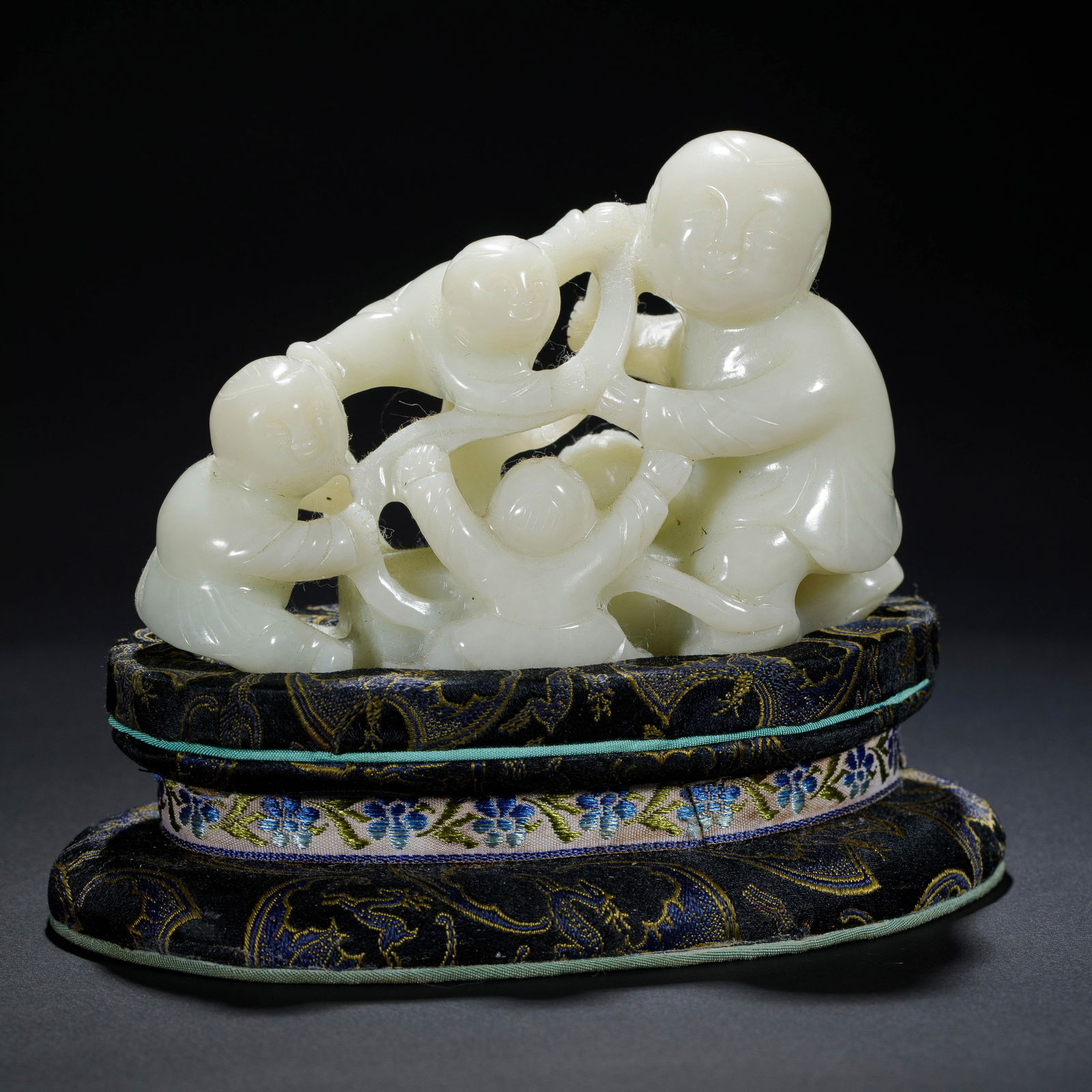 A FINECARVED WHITE JADE BOYS GROUP: A FINECARVED WHITE JADE BOYS GROUP This charming group carving is crafted from a single piece of luminous, white Hetian jade, depicting four playful boys in a lively tug-of-war. The composition is