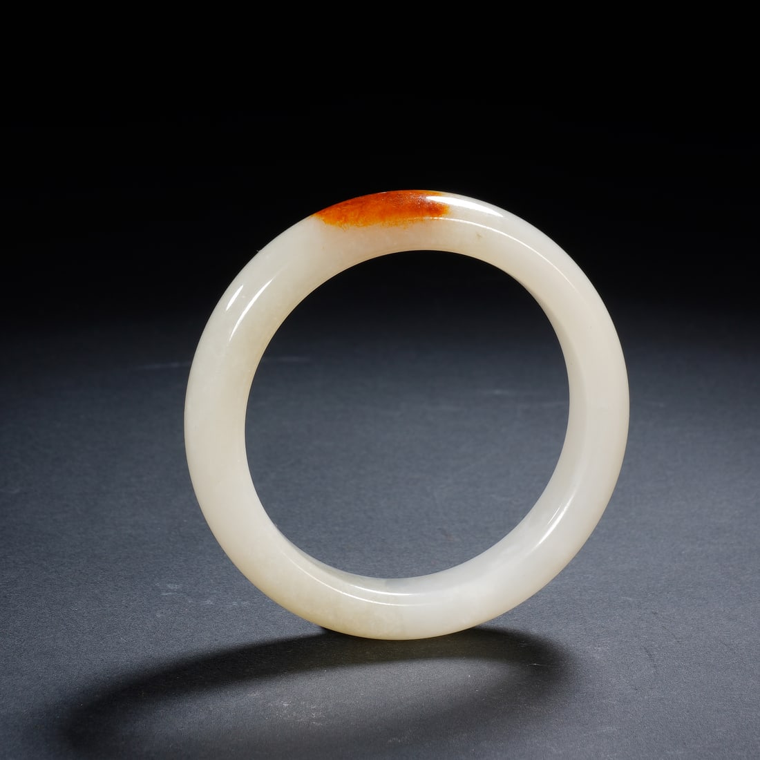 A WHITE JADE BANGLE: A WHITE JADE BANGLE An elegant Chinese bangle, carved from a single piece of smooth, milky-white Hetian jade, distinguished by a striking patch of natural russet skin along its upper curve. The warm,