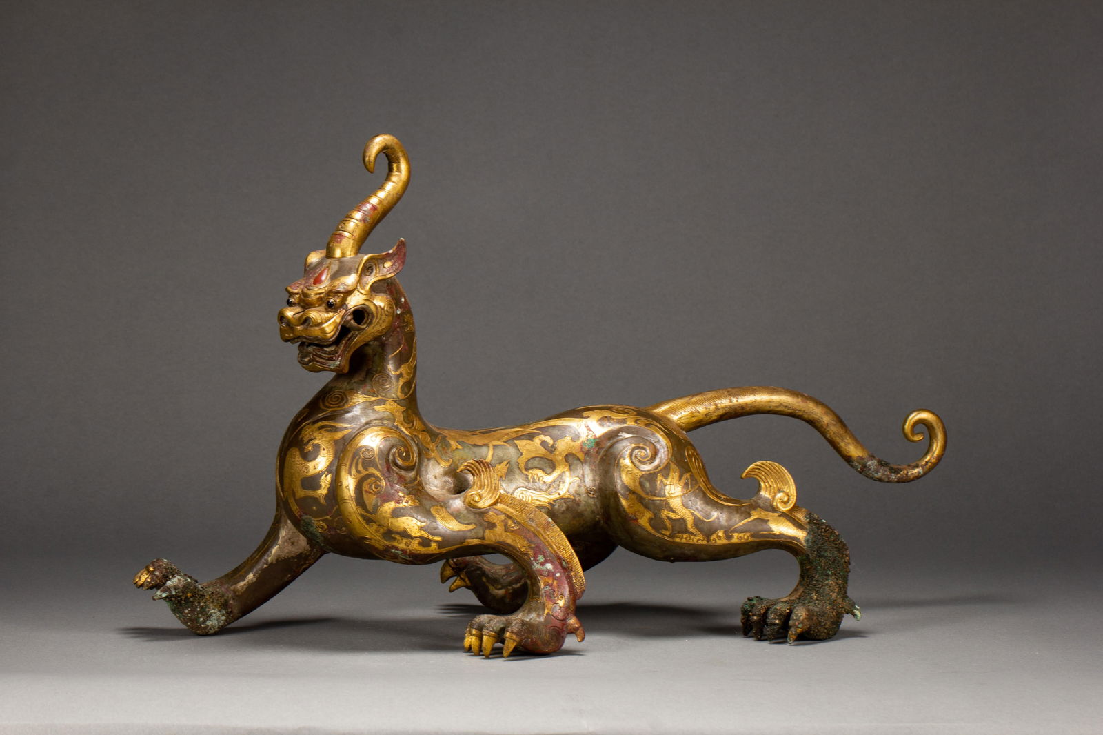 A GOLD AND SILVER INLAID BRONZE MYTHICAL BEAST: A GOLD AND SILVER INLAID BRONZE MYTHICAL BEAST A Chinese gold-inlaid bronze mythical beast, depicted in a dynamic striding pose with a single spiraling horn, open jaws, and a curled tail. The bronze