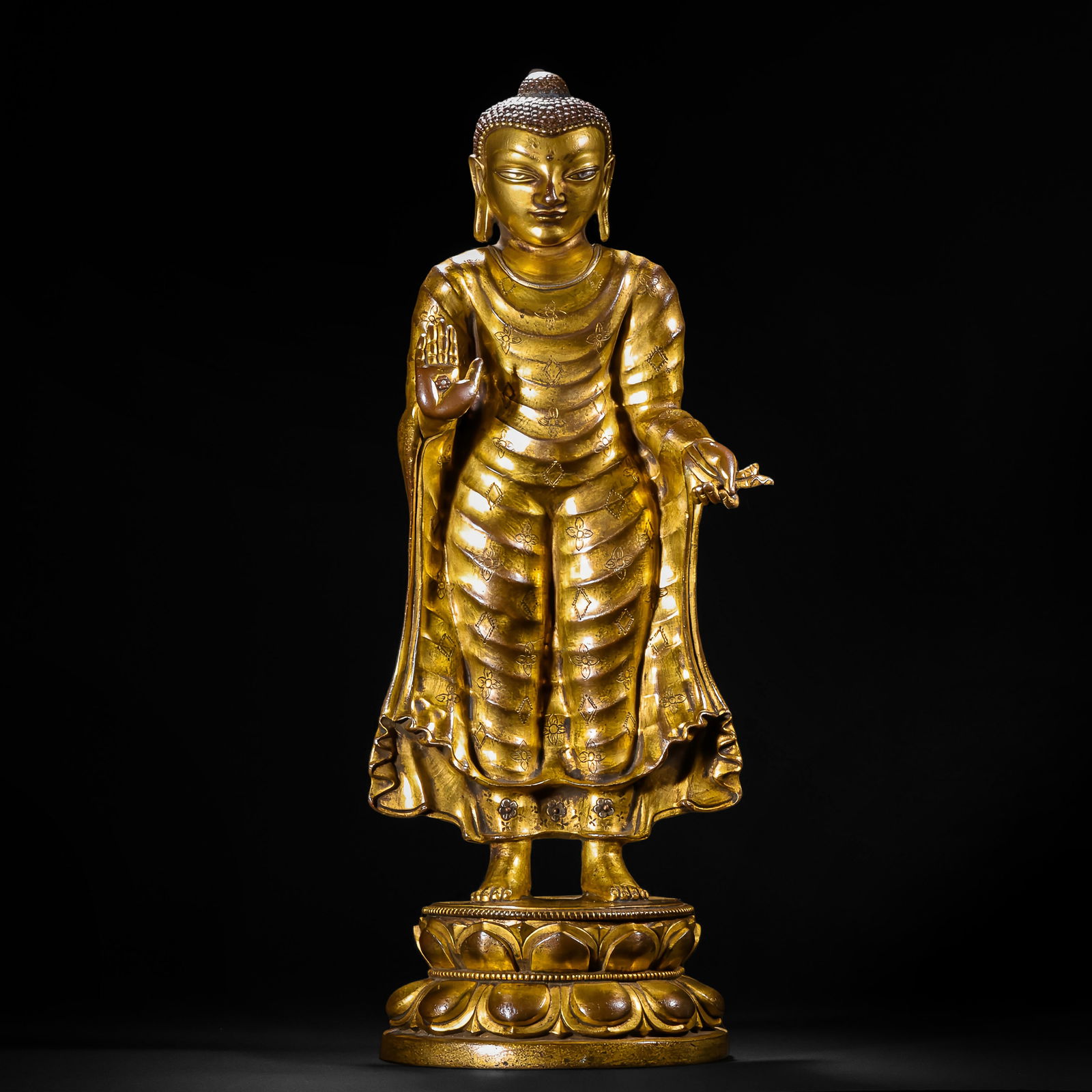 A BRONZE-GILT STANDING SHAKYAMUNI: A BRONZE-GILT STANDING SHAKYAMUNI A Chinese gilt-bronze standing Buddha statue, depicted with a serene expression, right hand in abhaya mudra and left hand holding a small sprig. The figure wears a fl