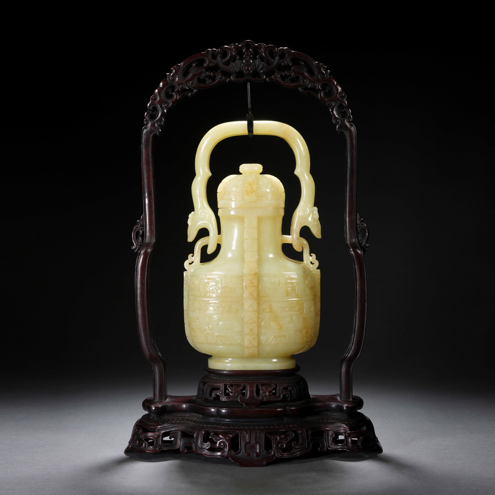 A RARE CARVED YELLOW JADE VASE WITH COVER: A RARE CARVED YELLOW JADE VASE WITH COVER An exceptional Chinese yellow jade archaistic ewer and cover, raised on an elaborate carved hongmu stand with a hanging arch frame. The compressed globular bo