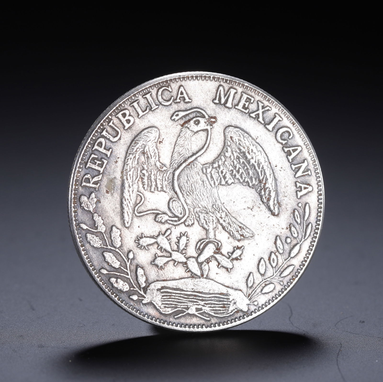 A MEXICAN SILVER COIN: A MEXICAN SILVER COIN A Mexican silver coin, struck with the iconic national emblem: an eagle clutching a serpent in its beak, perched atop a cactus, surrounded by oak and laurel branches. The legend