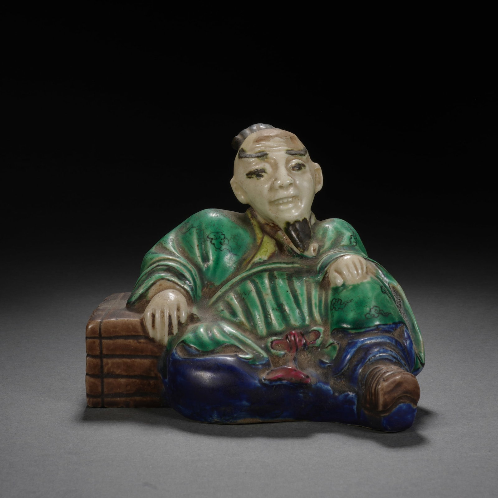 Chinese Famille Verte Glazed Seated Scholar Figure: Chinese Famille Verte Glazed Seated Scholar Figure This charming glazed ceramic figure depicts a reclining scholar, leaning casually against a stack of books. The figure is vividly dressed in a green 