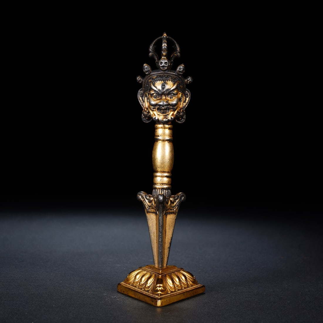 Chinese Gilt-Bronze Vajra Pestle (Phurba): Chinese Gilt-Bronze Vajra Pestle (Phurba) This finely crafted ritual phurba is cast in bronze with partial gilding, featuring a fierce Mahakala head at the apex adorned with skull ornaments, transitio
