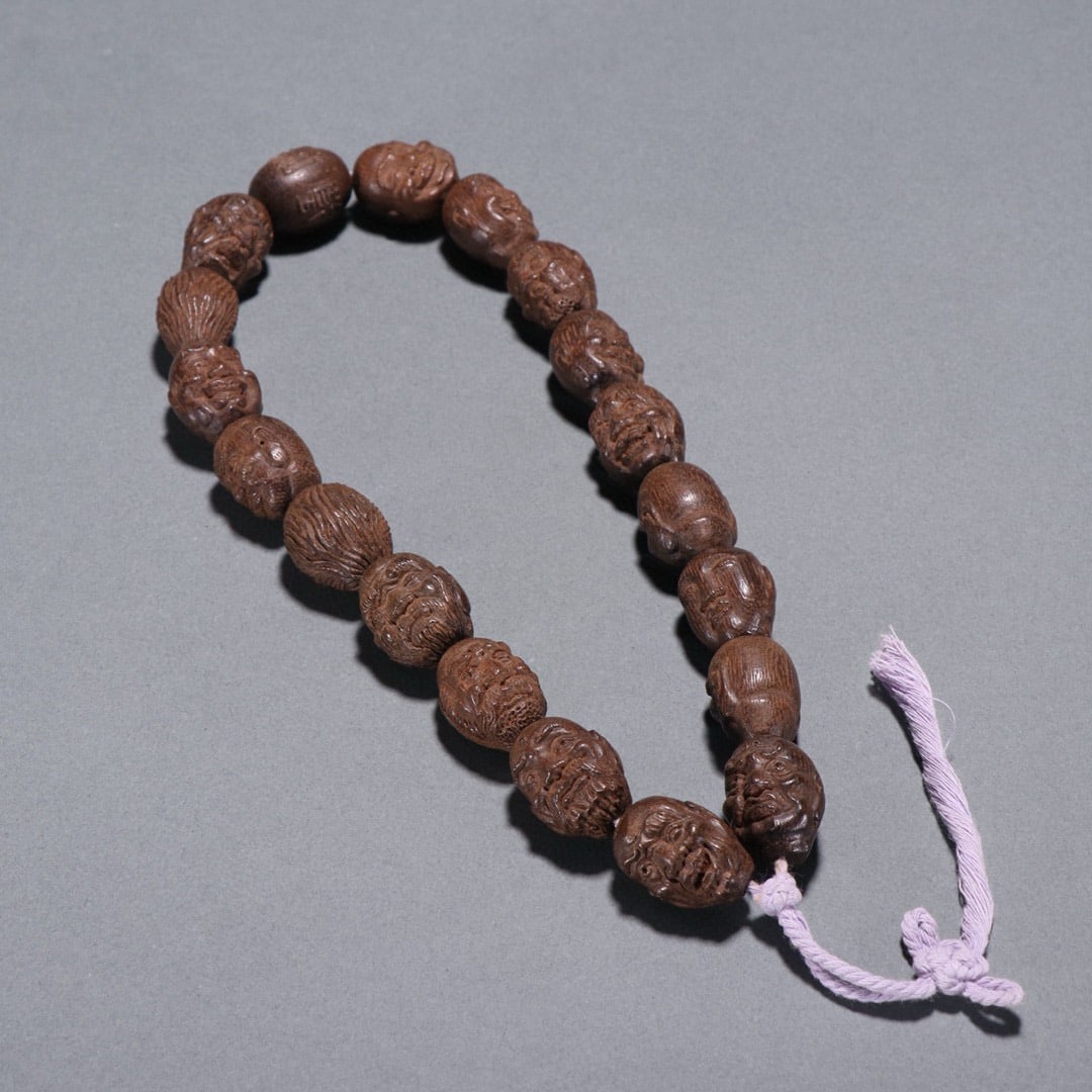 A CHINESE AGARWOOD PRAYER BEADS CARVED WITH LUOHAN HEADS: A CHINESE AGARWOOD PRAYER BEADS CARVED WITH LUOHAN HEADS This handheld strand is crafted from fine natural agarwood with a rich dark brown tone and distinctive aromatic grain, weighing 65.8 grams in t