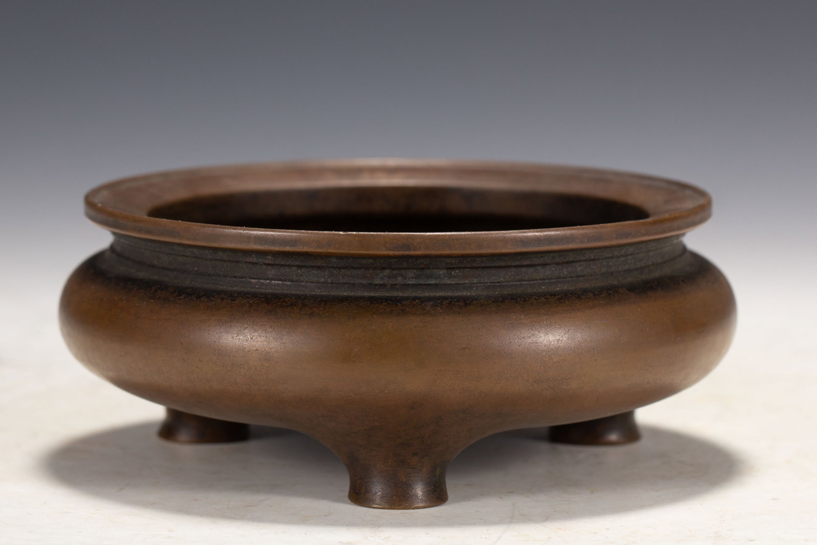 A CHINESE PLAIN BRONZE TRIPOD CENSER WITH RIBBED NECK: A CHINESE PLAIN BRONZE TRIPOD CENSER WITH RIBBED NECK This classic bronze censer features a wide flat flared rim above a narrow neck adorned with fine concentric horizontal ribs. The body swells into 