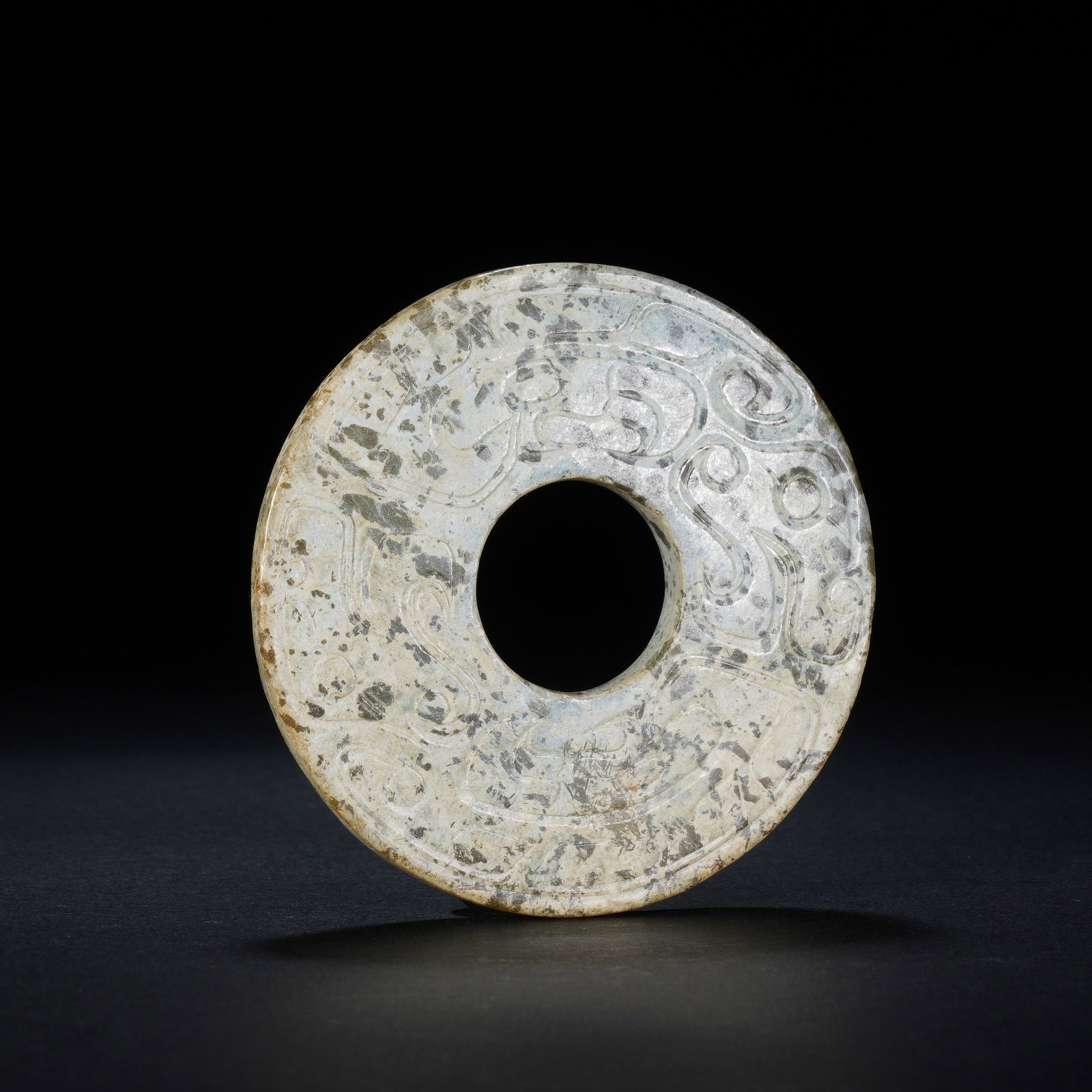 A CHINESE ARCHAIC CARVED JADE BI DISC: A CHINESE ARCHAIC CARVED JADE BI DISC This single circular jade bi disc measures 7.5 cm in diameter and weighs 71 grams. The flat, perforated body is intricately carved with winding stylized beast