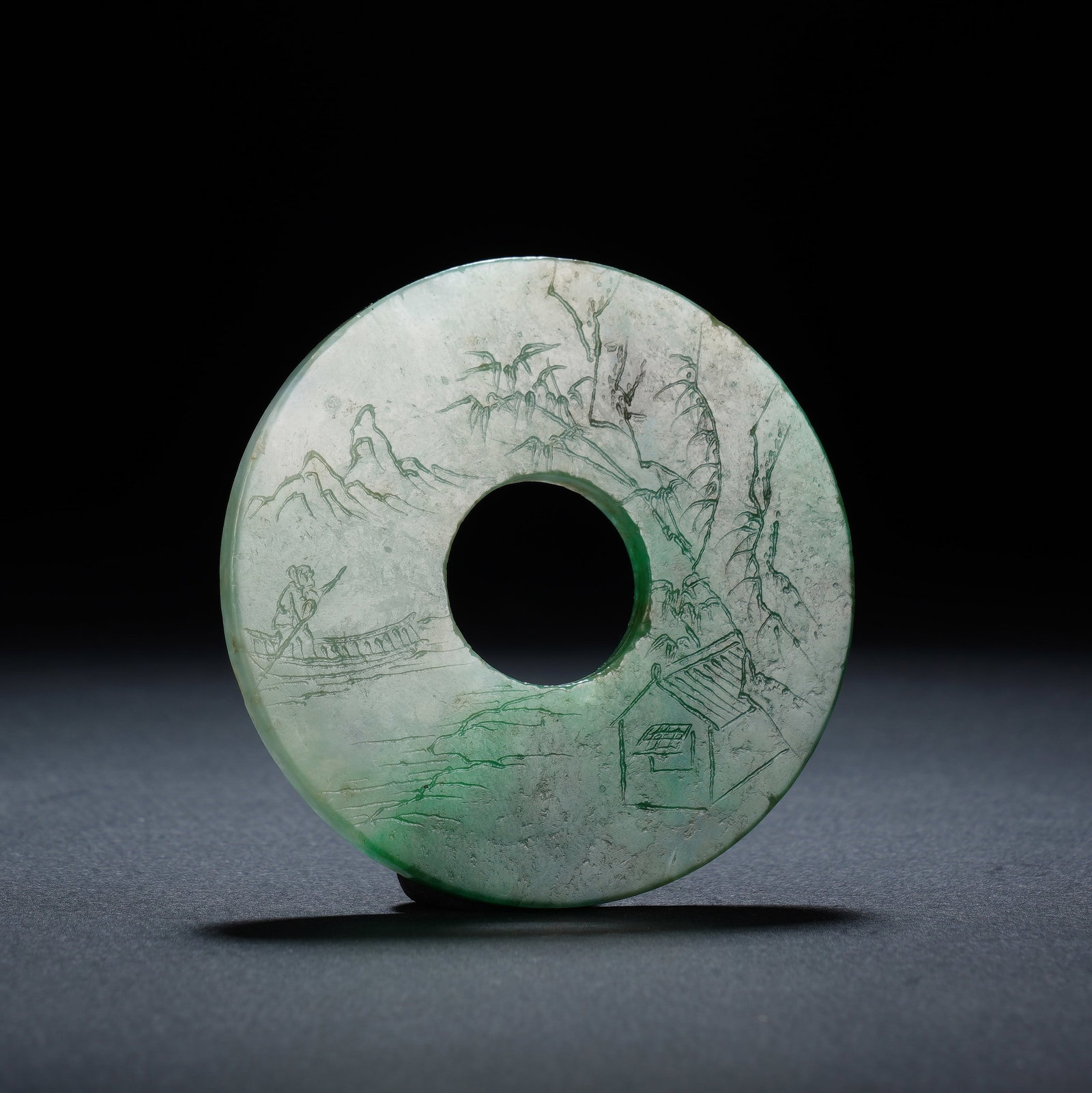 A CHINESE ENGRAVED JADEITE BI DISC: A CHINESE ENGRAVED JADEITE BI DISC This circular jadeite bi ornament measures 5.8 cm in diameter and 0.4 cm thick, weighing 34 grams. The translucent pale jadeite is naturally suffused with soft emera