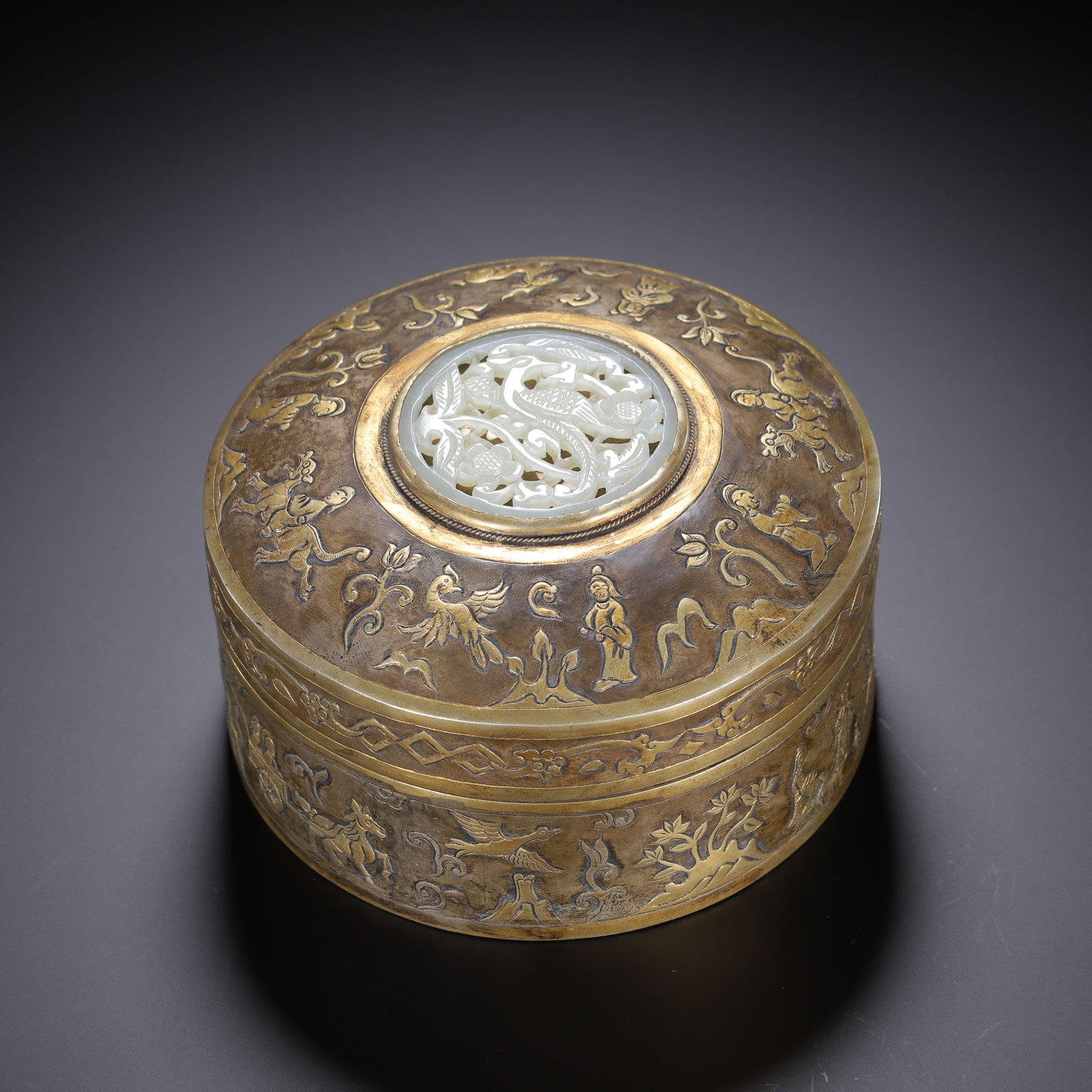 A CHINESE GILT BRONZE JADE-INSET COVERED BOX: A CHINESE GILT BRONZE JADE-INSET COVERED BOX A circular lidded box crafted from bronze with partial gilding over a muted patinated base. The domed lid is centered with an openwork carved pale jade pla