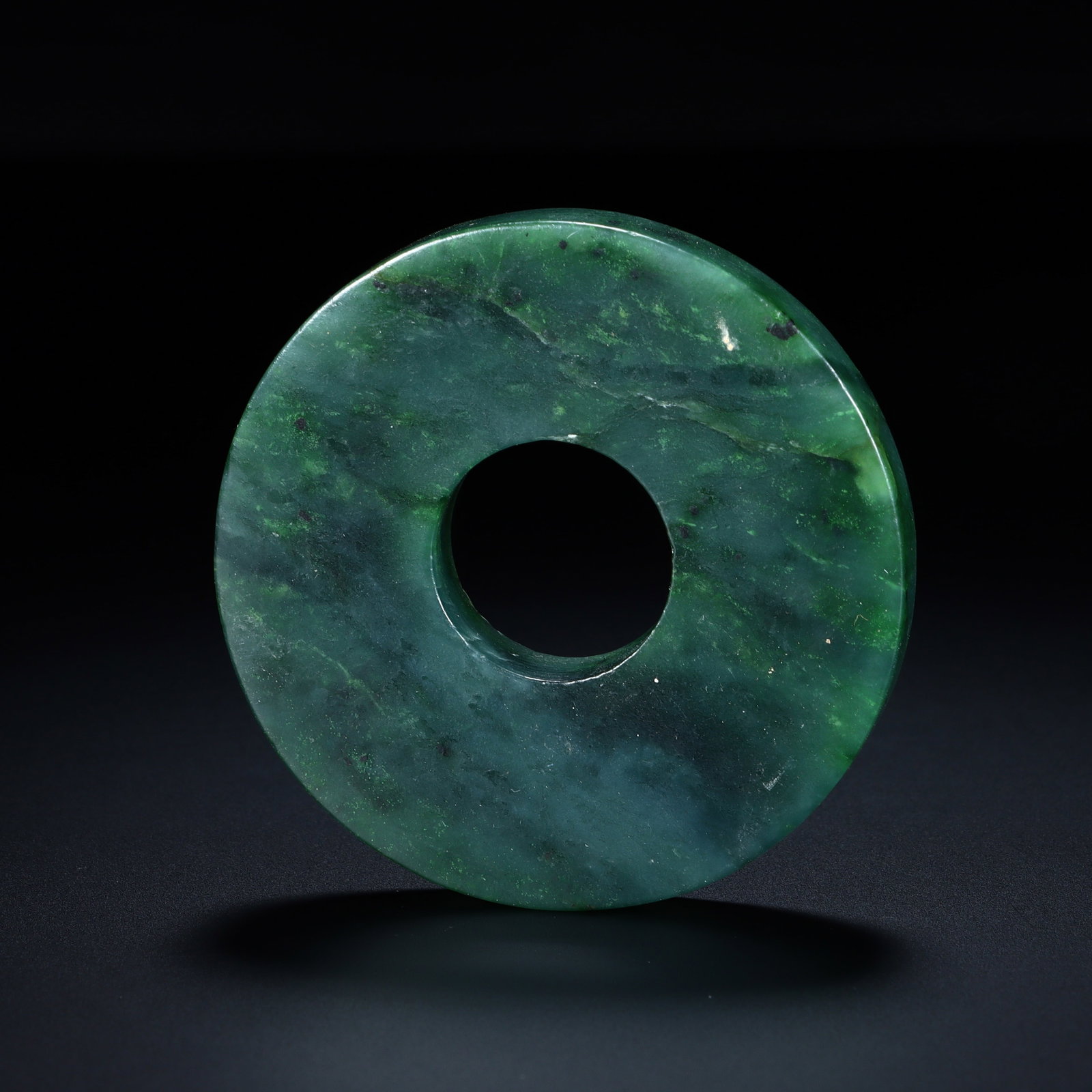 A CHINESE SPINACH GREEN JADE BI DISC: A CHINESE SPINACH GREEN JADE BI DISC This circular ritual bi disc is polished from solid nephrite spinach green jade with natural mottled deep green tonalities scattered with fine dark mineral speckle