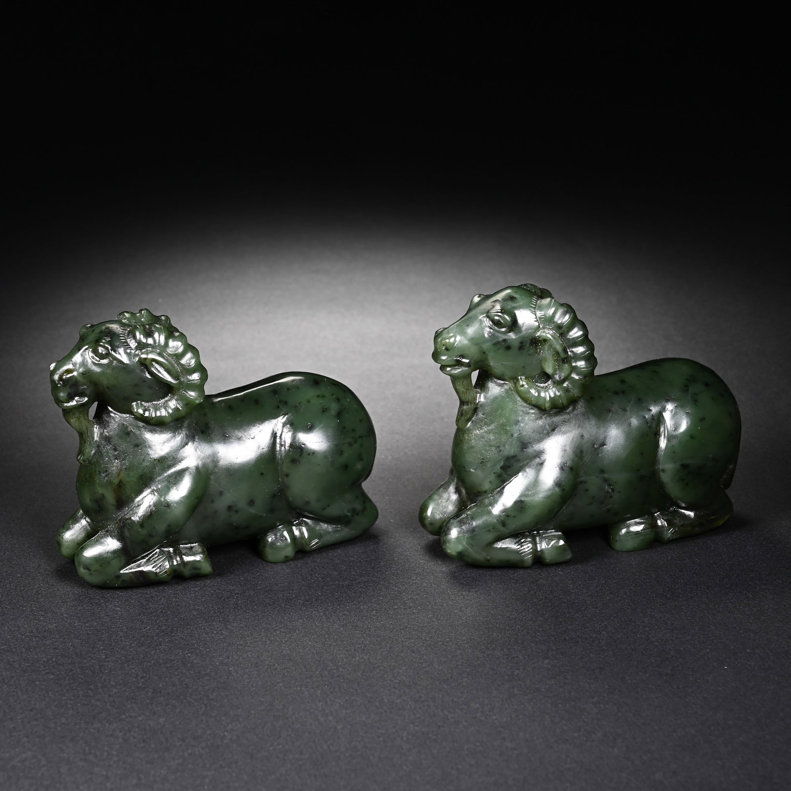 A PAIR OF CHINESE SPINACH GREEN JADE RECUMBENT RAMS: A PAIR OF CHINESE SPINACH GREEN JADE RECUMBENT RAMS This matching pair of animal ornaments is carved from natural spinach green Hetian jasper jade scattered with characteristic dark mineral speckles, 