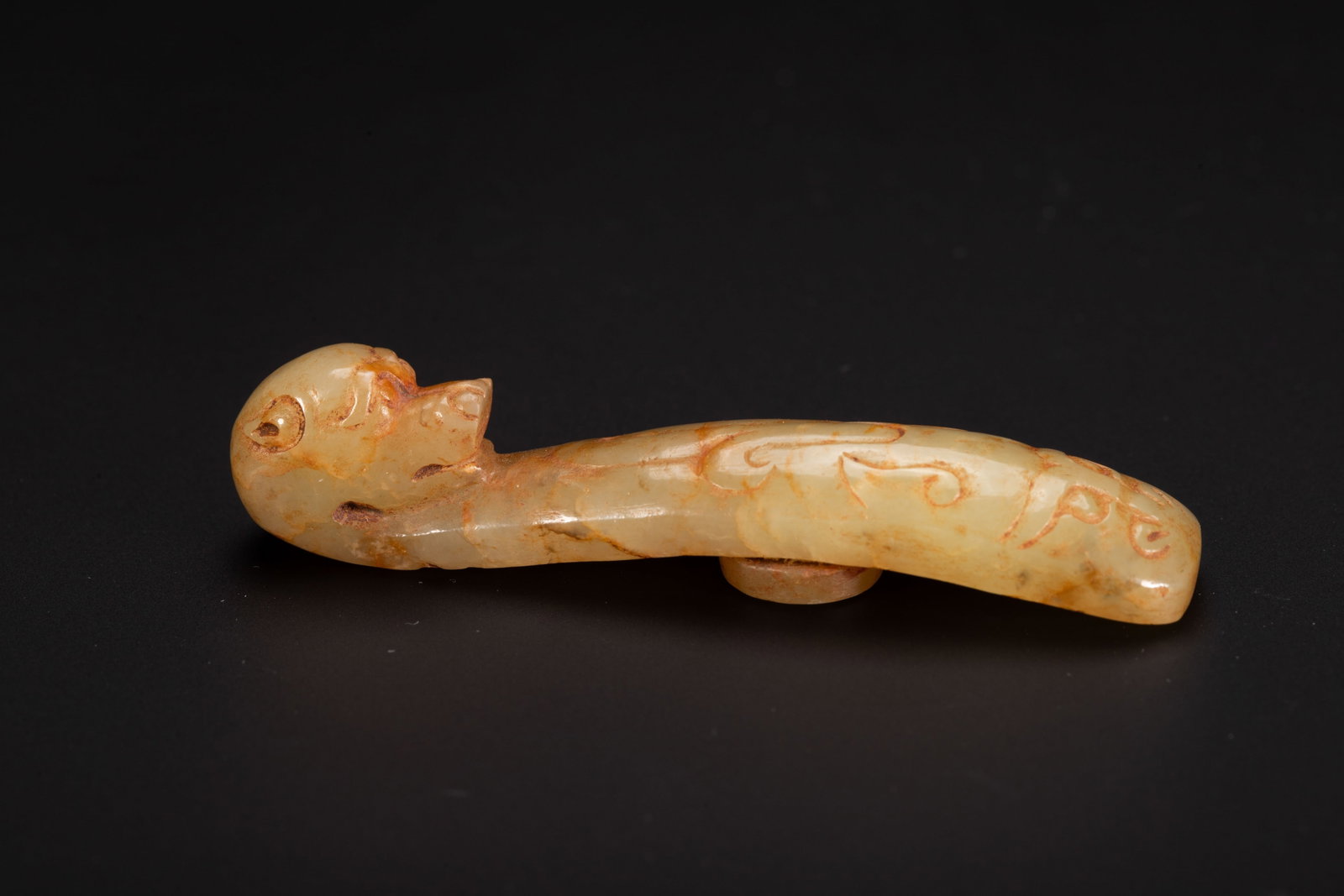 A CHINESE HETIAN JADE DRAGON BELT HOOK: A CHINESE HETIAN JADE DRAGON BELT HOOK This slender garment fastener is carved from pale creamy Hetian nephrite jade naturally streaked with warm russet inclusions. The terminal is finely shaped as a 