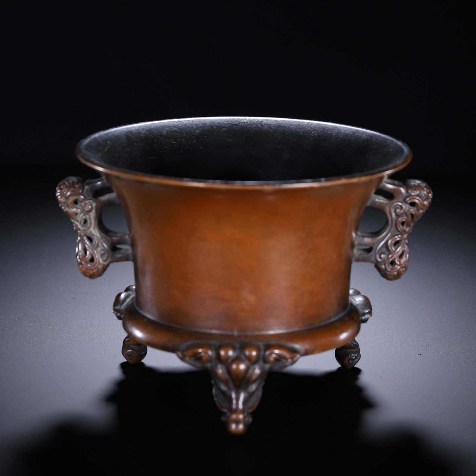 A CHINESE BRONZE TRIPOD CENSER: A CHINESE BRONZE TRIPOD CENSER A flared cylindrical censer cast from solid bronze with smooth warm reddish-brown patina and dark polished interior. Ornate openwork scroll-form handles extend symmetric