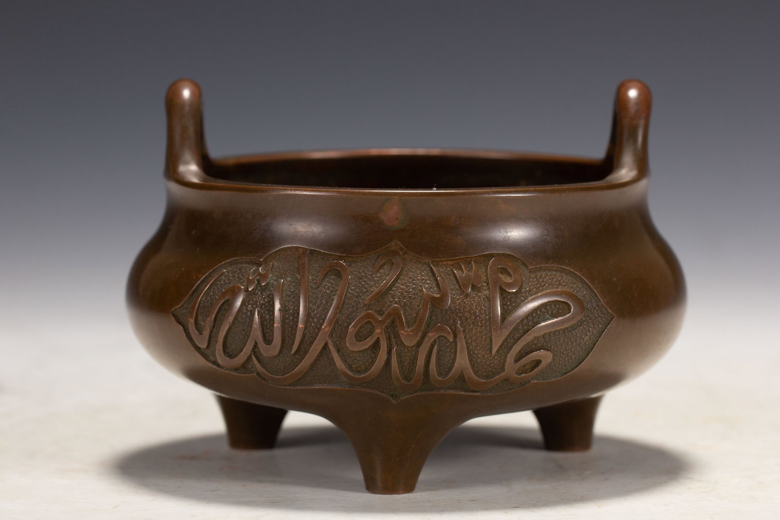 A CHINESE BRONZE TRIPOD CENSER WITH ARABIC SCRIPT CARTOUCHE: A CHINESE BRONZE TRIPOD CENSER WITH ARABIC SCRIPT CARTOUCHE This rounded bronze censer rests upon three tapered conical supports and features a pair of upright cylindrical loop handles rising from its