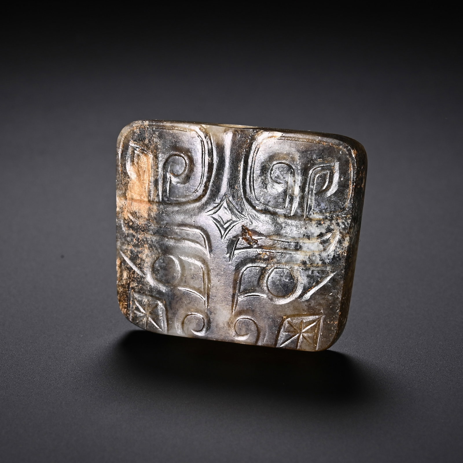 A CHINESE JADE TAOTIE MASK PENDANT: A CHINESE JADE TAOTIE MASK PENDANT This rounded rectangular pendant is carved from Hetian jade permeated with mottled grey, black and russet earth patina from prolonged burial. Its front surface bears