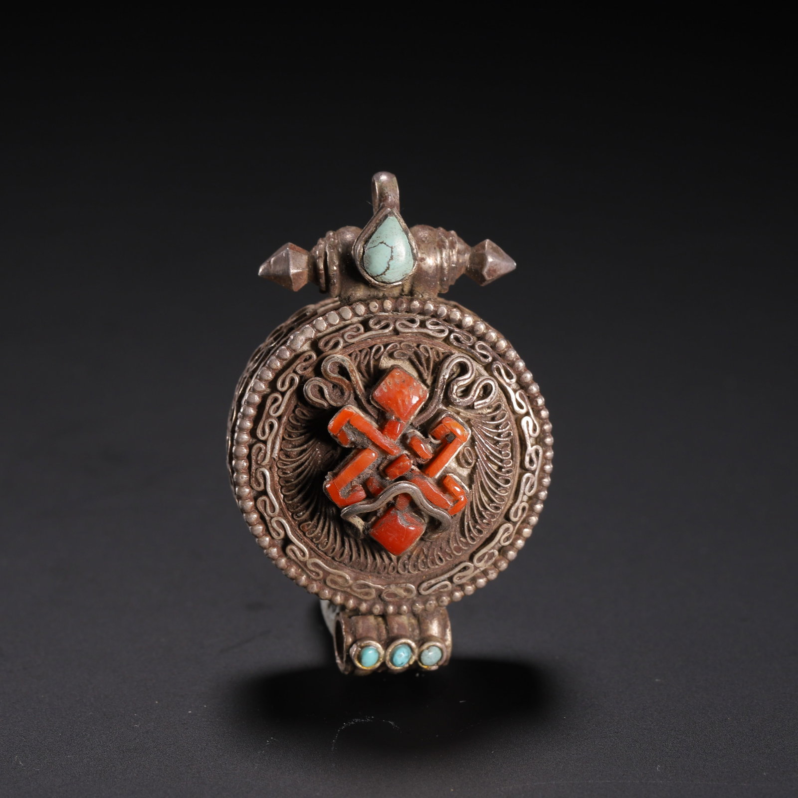 A CHINESE TIBETAN INLAID SILVER GAU BOX: A CHINESE TIBETAN INLAID SILVER GAU BOX This miniature circular Tibetan gau pendant is wrought from aged silver, fitted with a suspension loop for neck wear. Its front face features filigree scrollwor