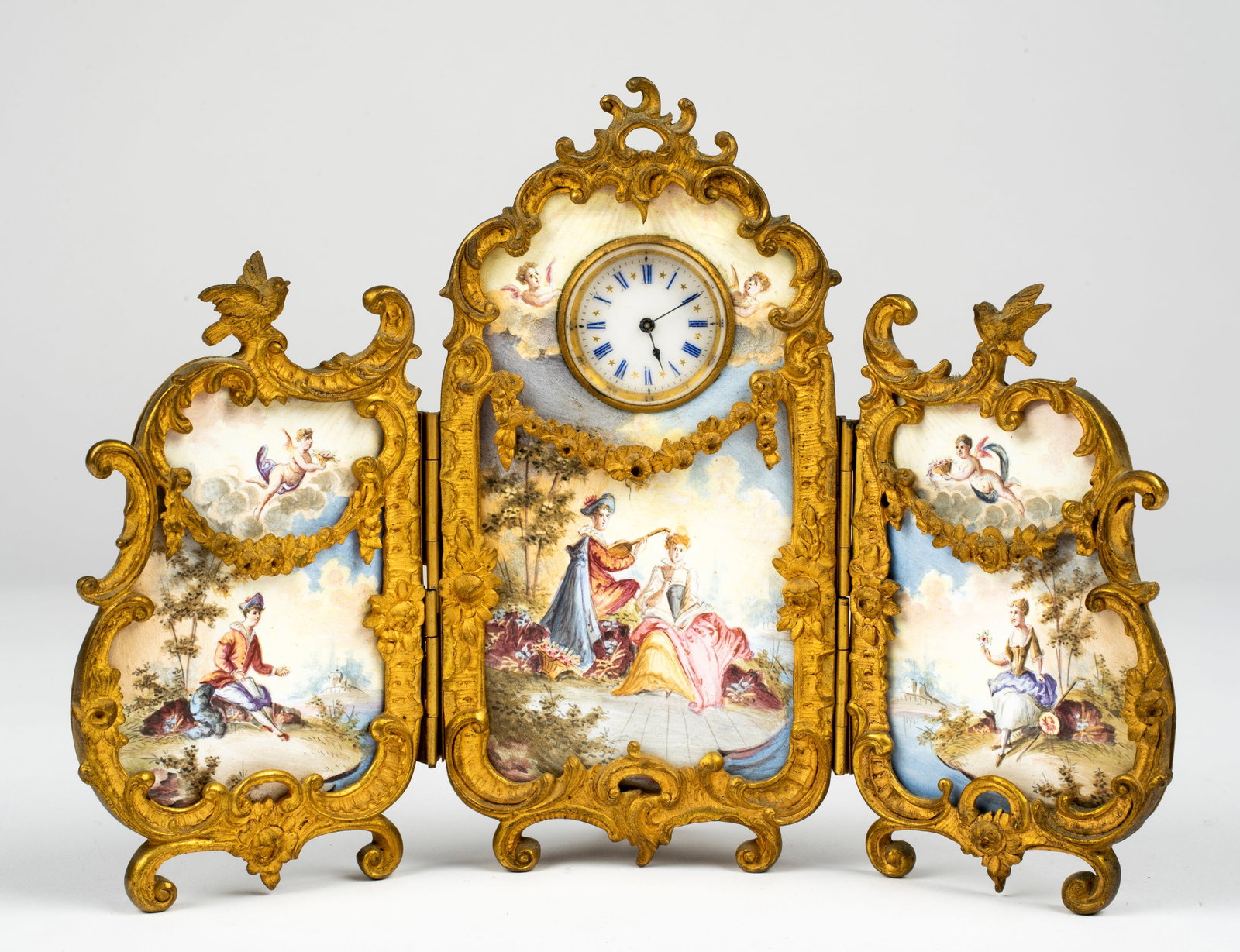 A French or Austrian Gilt and Enamel Table Screen and Clock: Three hinged sections each with enamel Continental landscape scenes with four figures total, the side panels each with a hovering cherub and the center an inset clock with blue roman numerals and gold