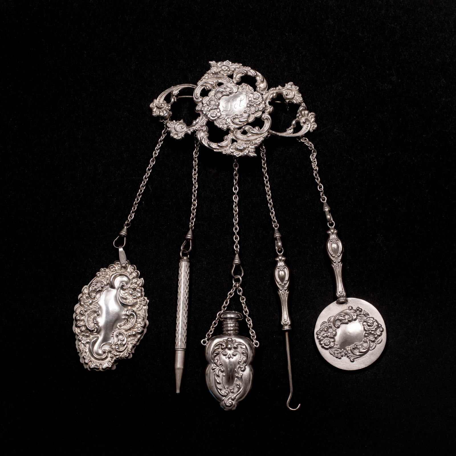 An Antique Sterling Silver Repousse Chatelaine: Marked "Sterling", large folate and scroll decorated brooch with "Dot" engraved in the center panel with five attachments each connected with a dog clip, a writing pad with four bone pages, a pencil, 