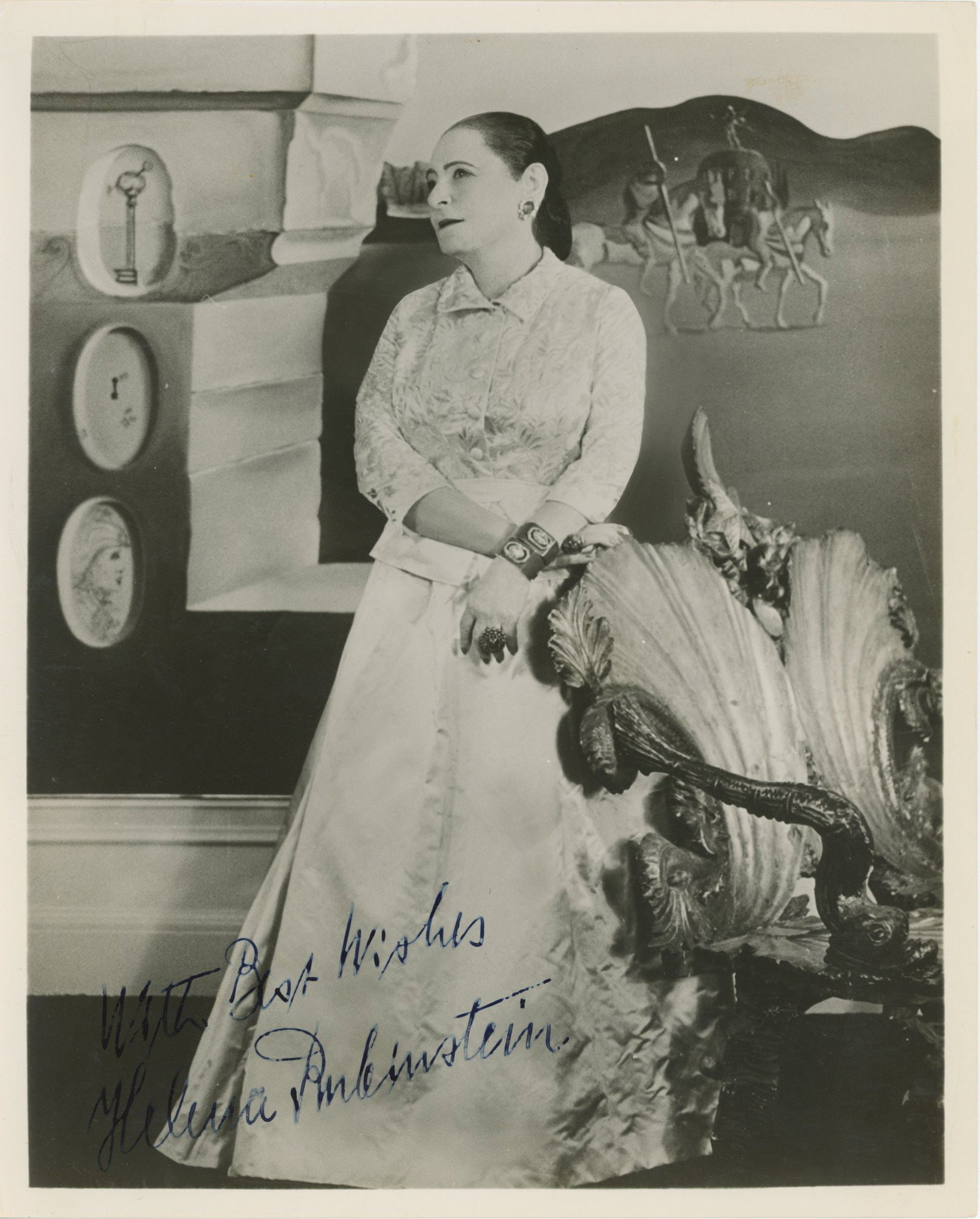 Helena Rubinstein – Cosmetics Pioneer – Autographed 8 x 10 Photograph – Inscribed: Details: Helena Rubinstein autographed 8 x 10” glossy photograph depicting the renowned cosmetics entrepreneur posed in an elegant interior setting beside a decorative sculptural chair. The