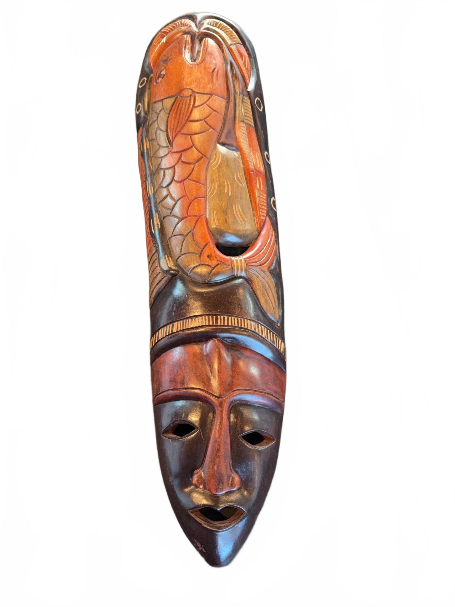 Hand-Carved Wooden Tribal Mask: Carved wooden tribal mask depicting a stylized face with an intricately detailed fish motif above the brow. The mask features multitone staining and incised designs accentuating both facial features