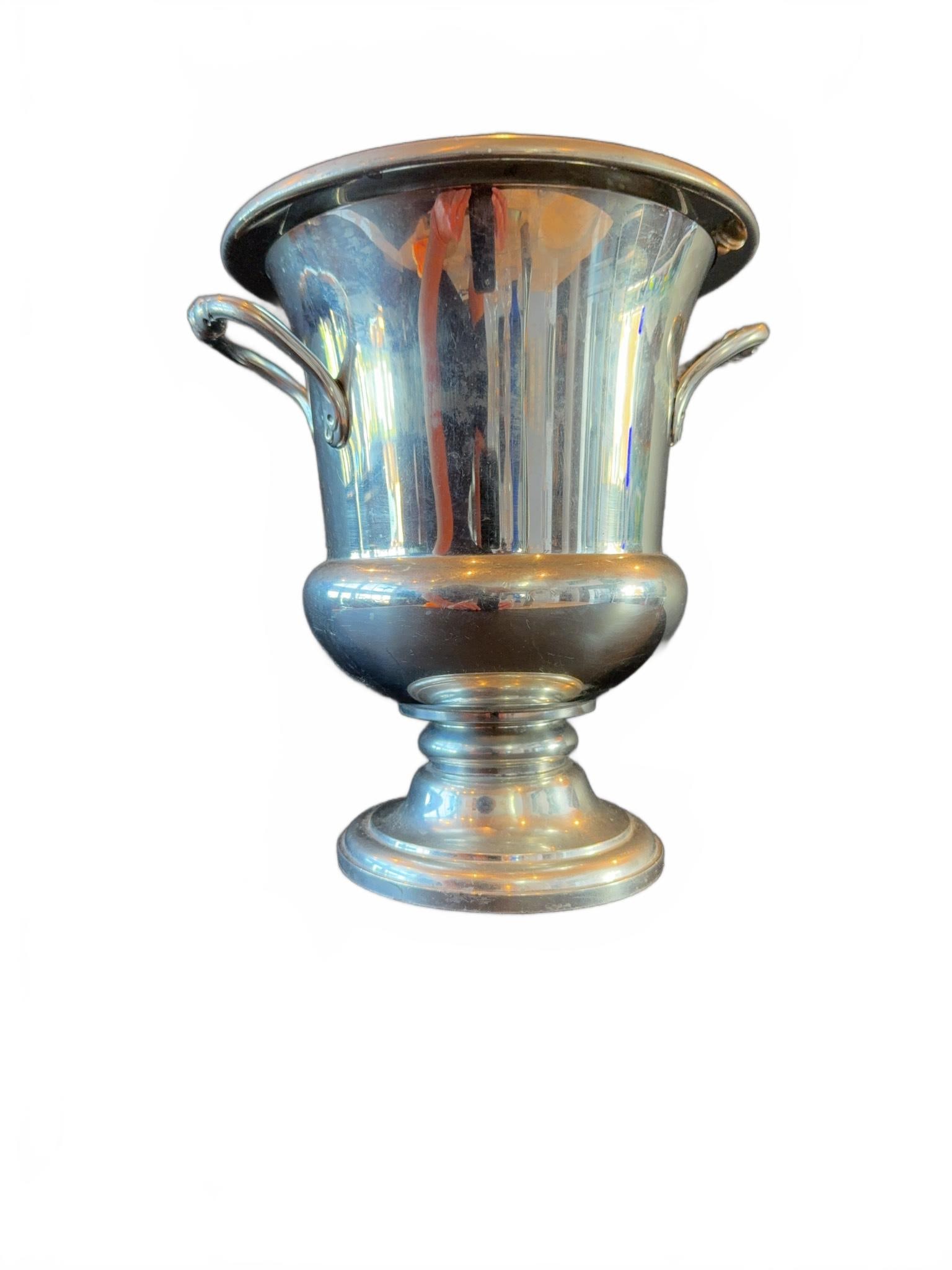 Silver Plate Wine Cooler with Dual Handles: Silver plate wine cooler featuring two ornate side handles and a pedestal base. The piece has a flared rim and a smooth, reflective finish, showing some surface wear consistent with regular use.