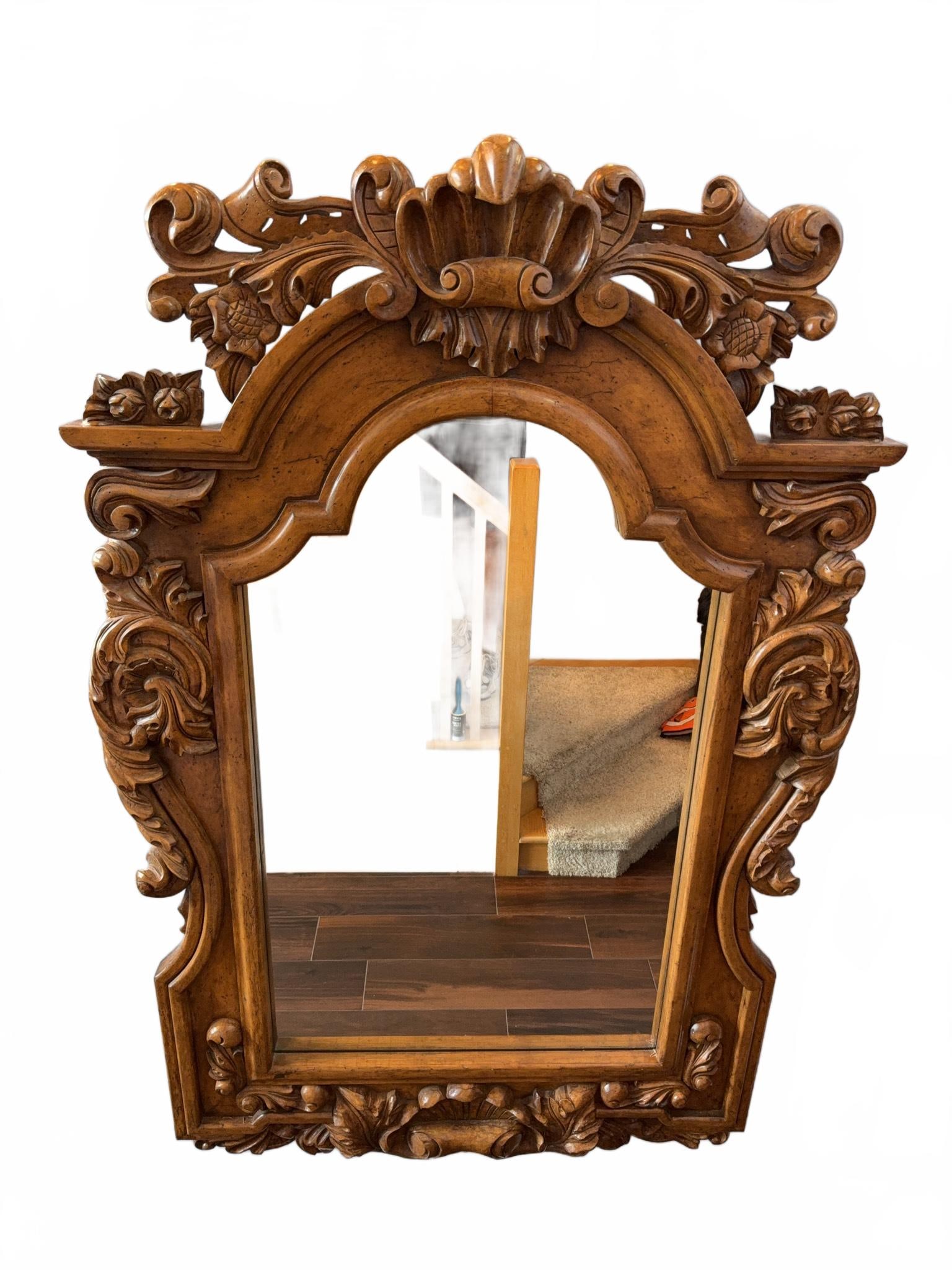Ornate Hand-Carved Wood Wall Mirror: Large wall mirror featuring an elaborate hand-carved wood frame with floral and shell motifs. Warm natural wood finish, detailed craftsmanship, and scrollwork throughout. Rectangular with arched top d