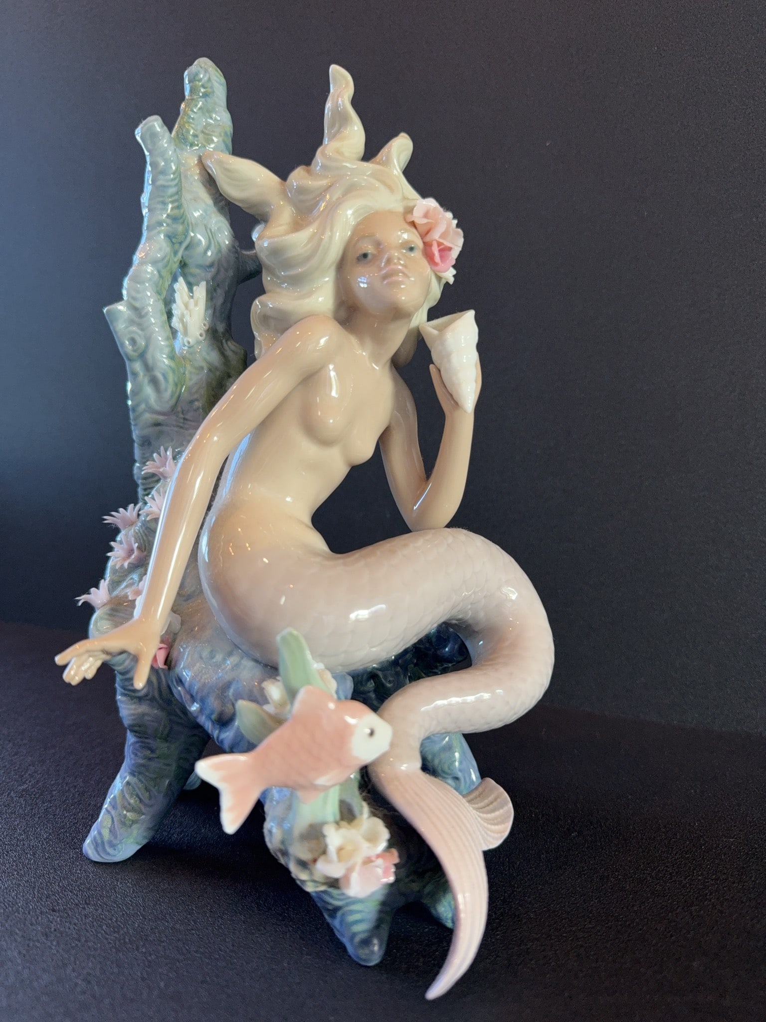 Lladro Porcelain Mermaid "Ocean Beauty" Figurine with Coral: Lladro Ocean Beauty Mermaid porcelain figurine depicting a mermaid seated on a coral formation, holding a conch shell. Features detailed pastel coloring, delicate flowers, and a fish at the base.