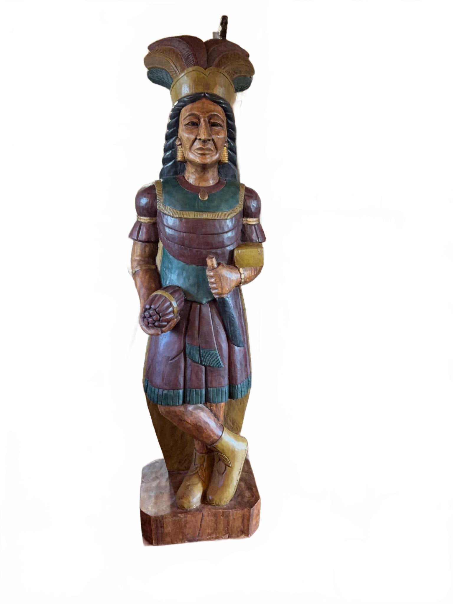5'11" Hand-Carved Wood Cigar Store Indian Figure: Polychrome hand-carved wood cigar store Indian figure featuring a feathered headdress, detailed facial features, crossed legs, and traditional attire. The figure holds a bundle of cigars, reflecting c