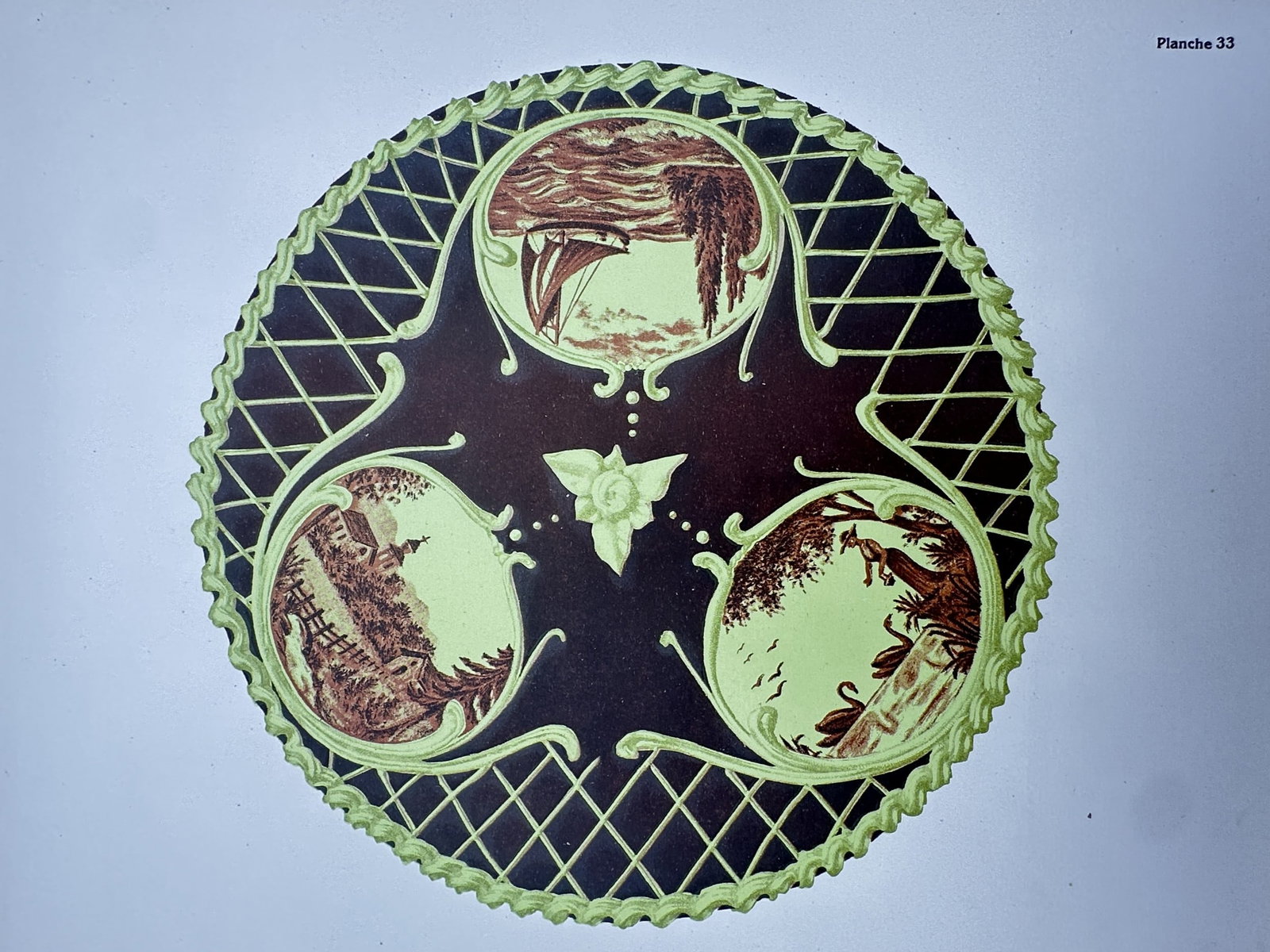 Thérèse de Dillmont (1846–1890), after – Circular Design with Three Tondo Landscapes –: "Planche 33" features a complex circular pattern containing three small tondo vignettes depicting a sailboat on a lake, a rural village, and a lakeside scene with swans. These pastoral scenes are inte