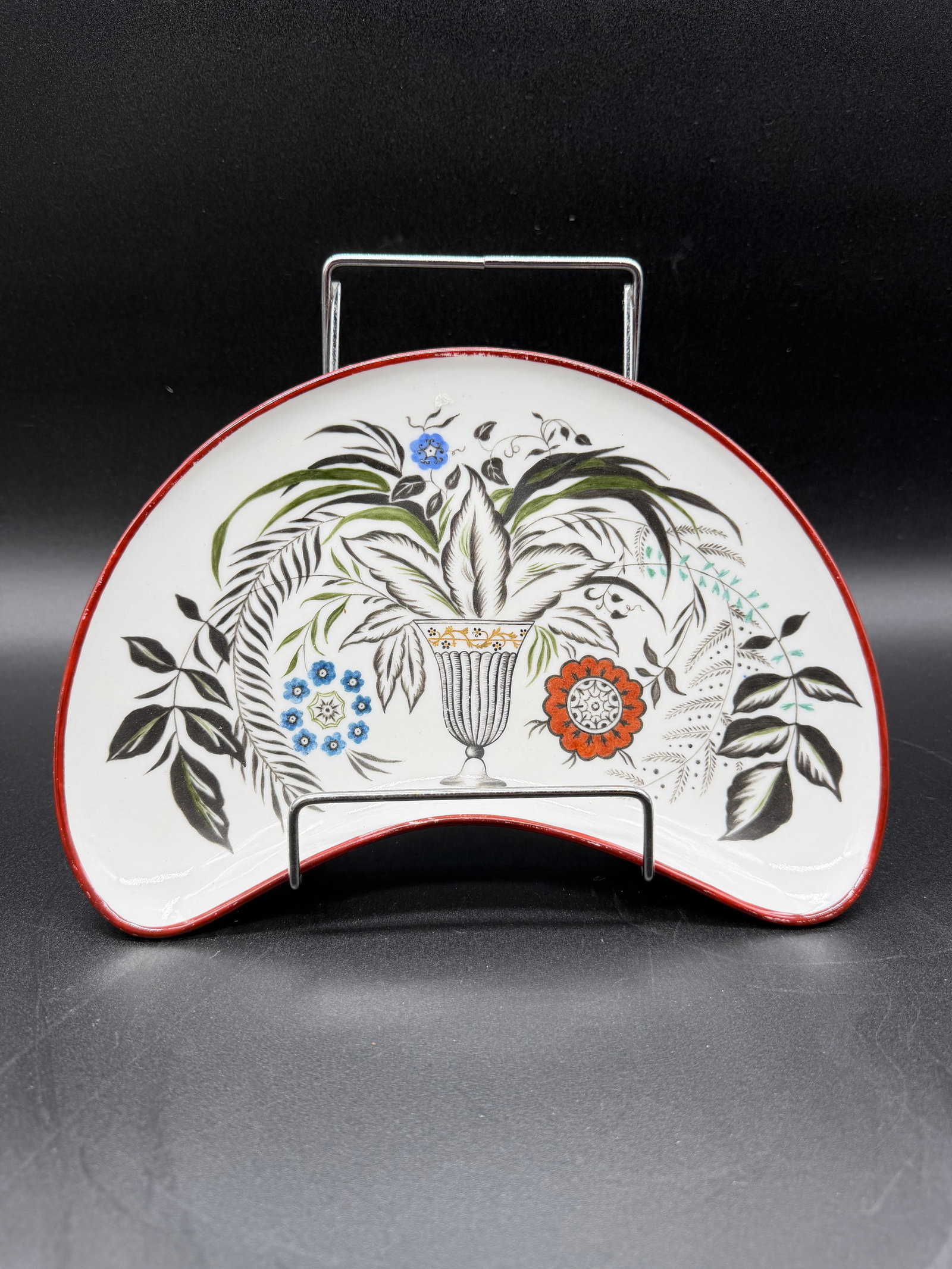 Sergei Vassilievich Chekhonin (1878–1936) | A Porcelain Side Dish, State Porcelain Manufactory,: Oval porcelain side dish with red rim, hand-painted in polychrome with a central vase filled with a leafy bouquet framed by stylised flowers on a white ground. Mark of the Imperial Porcelain Manufacto