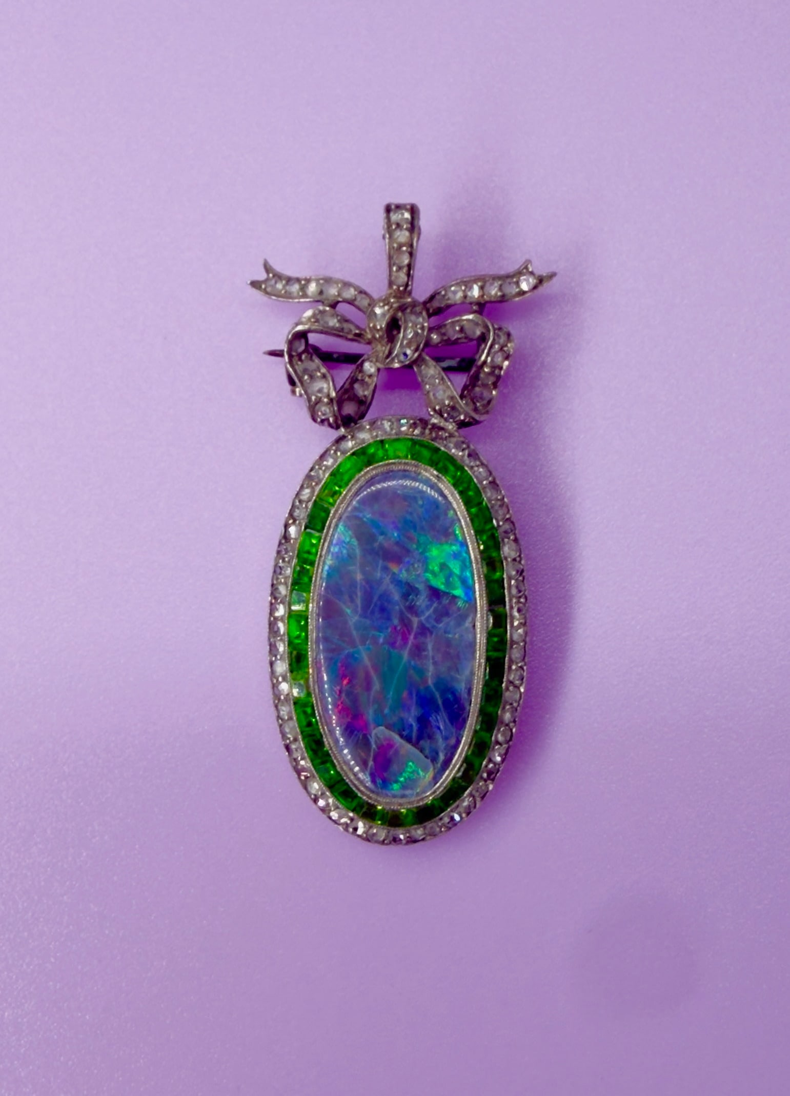 A Russian Black Opal, Emerald and Diamond Pendant-Brooch, circa 1900: Russian work, circa 1900Oval brooch?pendant in silver?coloured metal and gold, surmounted by a ribbon bow. It is set at the centre with an oval cabochon black opal displaying intense multicoloured pla