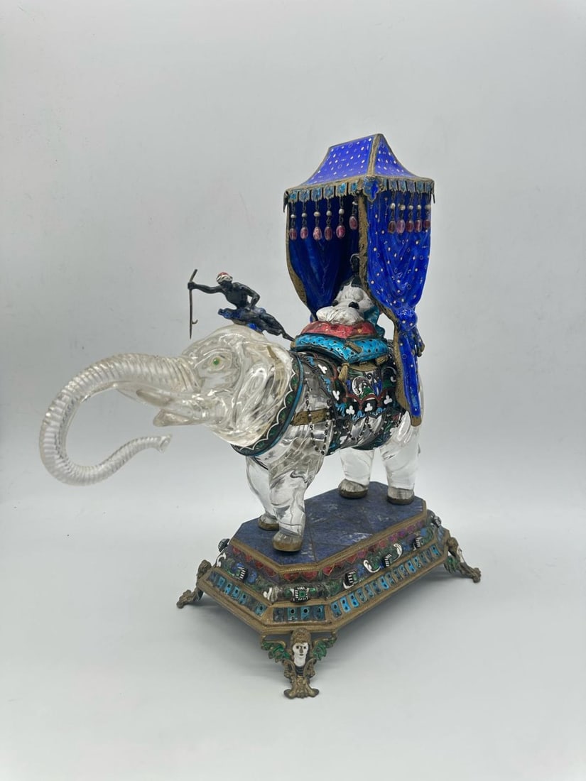ROCK CRYSTAL, CLOISONNÉ ENAMEL, AND LAPIS LAZULI CENTERPIECE DEPICTING AN ELEPHANT, LATE 19TH: An important centerpiece featuring a carved translucent rock crystal elephant with raised trunk and green stone-inlaid eyes. The figure is richly caparisoned with a gilt-metal or gilt-silver saddle, b