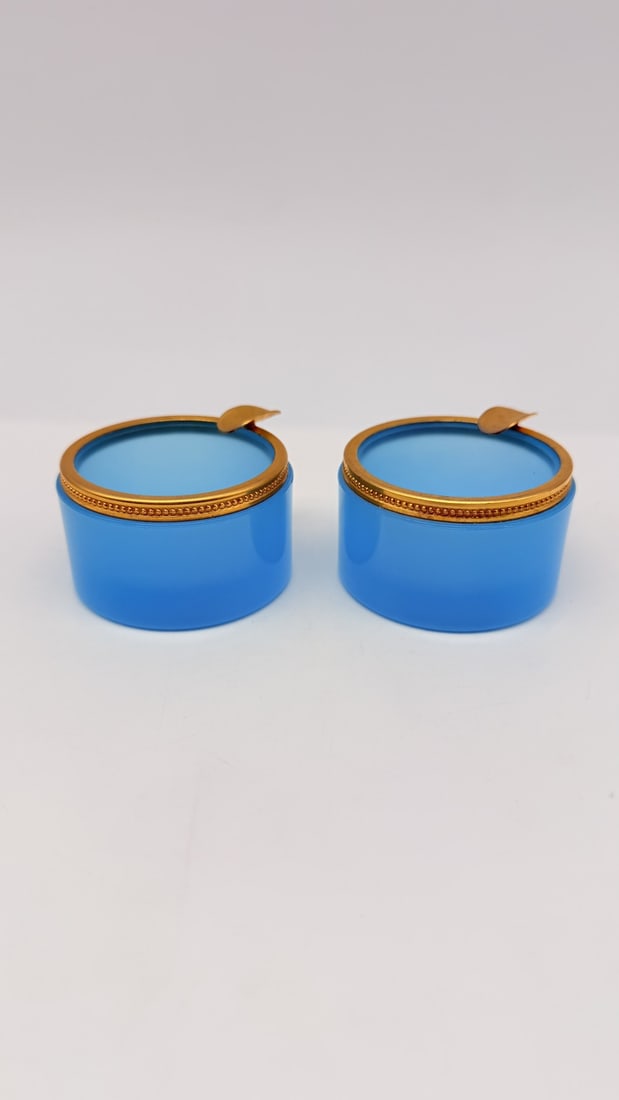 PAIR OF MURANO BLUE OPALINE GLASS ASHTRAYS WITH BRASS MOUNTS, MID-20TH CENTURY: A pair of small cylindrical ashtrays crafted in turquoise-blue opaline glass. Each piece features a smooth glass body fitted with a beaded brass metal rim incorporating a single cigarette rest.Italy (