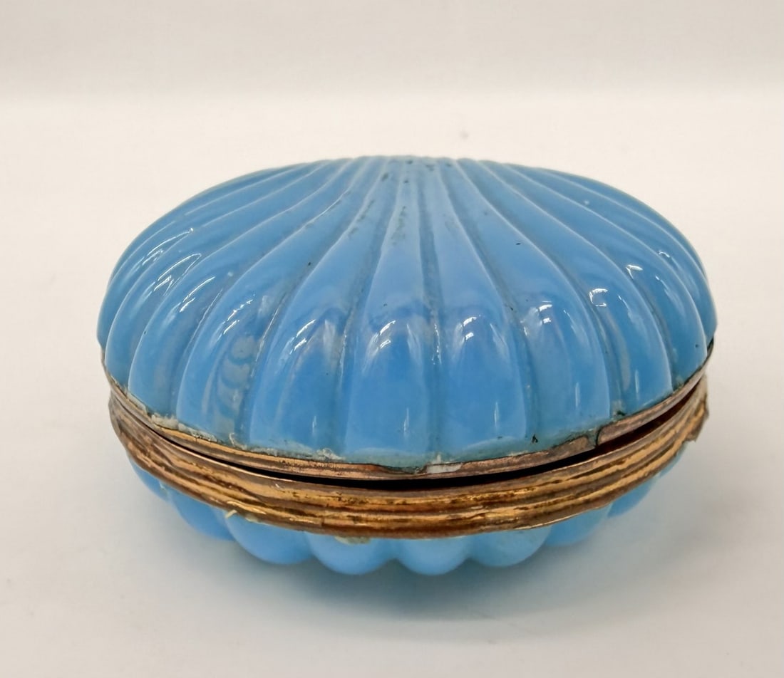 BLUE OPALINE GLASS BOX WITH BRONZE MOUNT, CHARLES X PERIOD, CIRCA 1820–1830: A miniature circular trinket box crafted in light blue opaline glass, known for its translucent soap-bubble effect. The cover and base are press-molded with radiating ribbed flutes. The two halves are