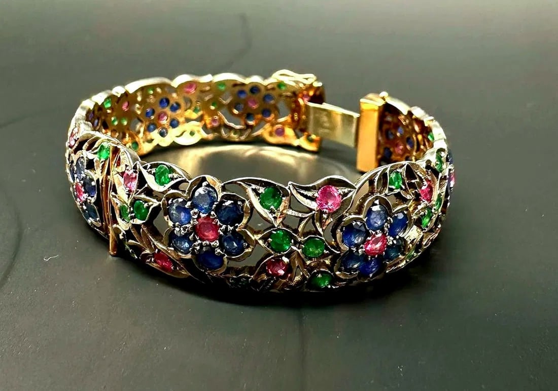 18K yellow gold and silver floral hinge bangle set with rubies, sapphires and emeralds: Important openable rigid bangle (hinged bangle) in 18K yellow gold (750/1000) and silver, featuring openwork, chased, and blackened scrollwork, foliage, and polychrome floral motifs. The outer side is