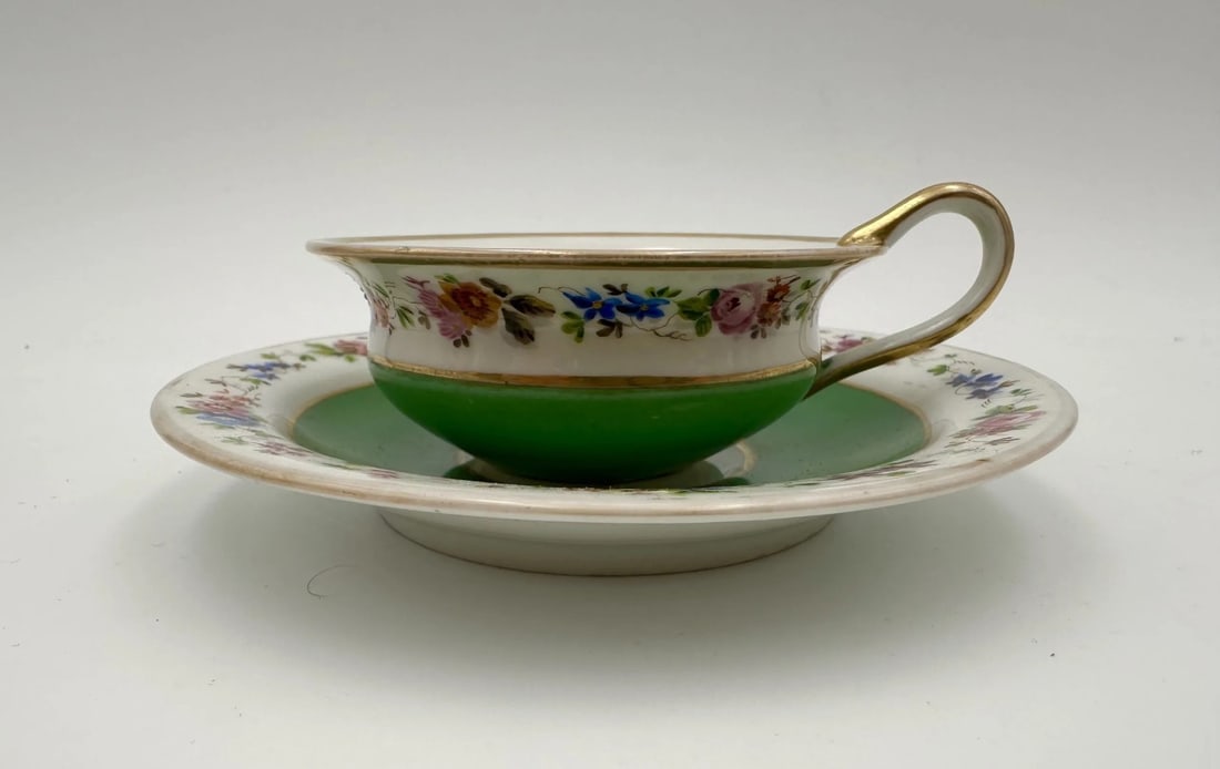 RUSSIAN IMPERIAL PORCELAIN CUP AND SAUCER, GARDNER FACTORY, 19TH CENTURY: A teacup and saucer set in hard-paste porcelain. The flared cup features an apple-green lower body and a gilded loop handle, while the upper white reserve is hand-painted with continuous polychrome fl