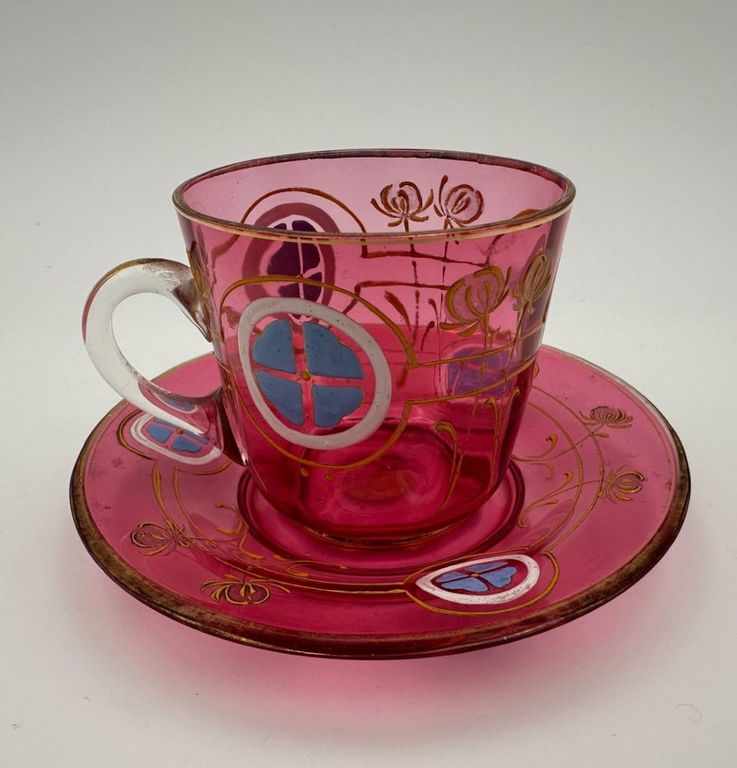 RUSSIAN IMPERIAL GLASS CUP AND SAUCER, ART NOUVEAU PERIOD, CIRCA 1900: A teacup and matching saucer set in translucent cranberry-pink crystal, featuring an applied clear glass C-scroll handle. The set is decorated with hand-painted Art Nouveau enamel work, incorporating 