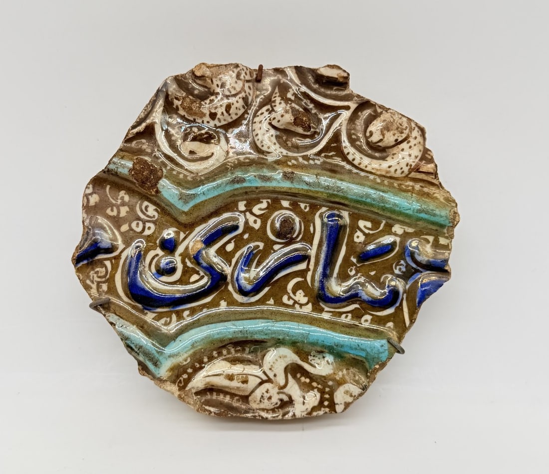 PERSIAN KASHAN CALLIGRAPHIC LUSTRE POTTERY TILE FRAGMENT, ILKHANID PERIOD, LATE 13TH CENTURY: A molded fritware (stonepaste) tile fragment with golden-brown lustre decoration, cobalt blue enamel, and turquoise highlights. The centerpiece features raised calligraphic script painted in cobalt bl