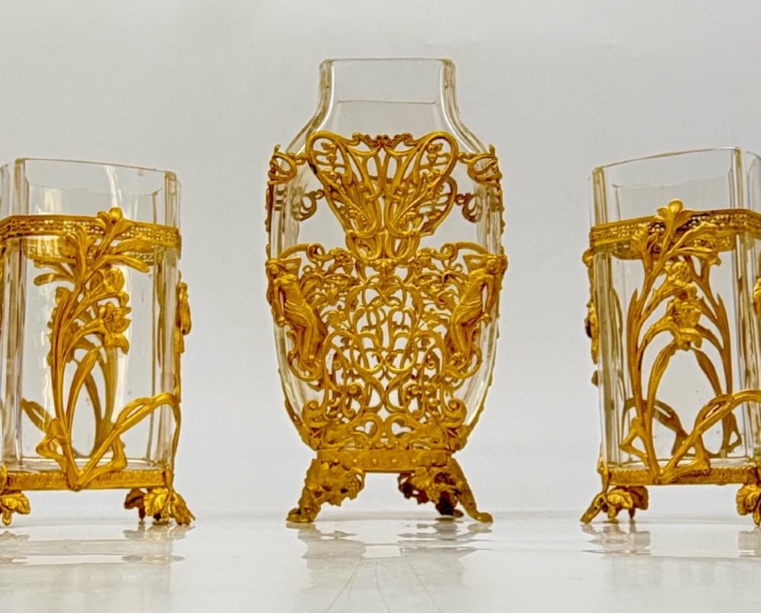 FRENCH ART NOUVEAU GILT-BRONZE MOUNTED CRYSTAL VASE GARNITURE SET, BACCARAT, CIRCA 1900: A three-piece Art Nouveau garniture set comprising a central vase and a pair of flanking side vases crafted in clear molded crystal. Each piece is encased in fine gilt-bronze (ormolu) openwork mounts 