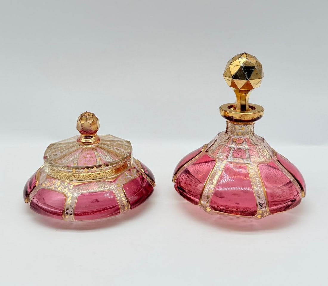 MOSER CABOCHON GLASS PERFUME BOTTLE AND COVERED BOX SET: Two-piece vanity set by Moser crafted from cranberry-pink and clear cut glass. The set includes a scent bottle with a faceted geometric gilt stopper and a matching low covered trinket box. Both pieces
